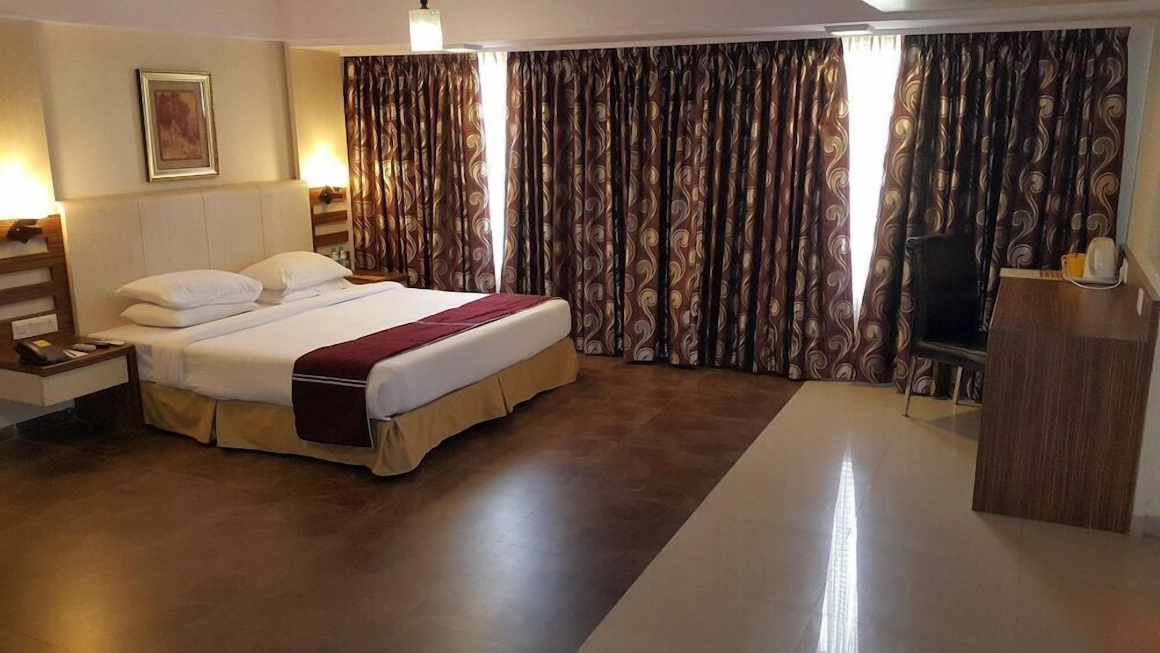 A hotel room with a bed, nightstand, lamp, curtains, and a desk with a chair.