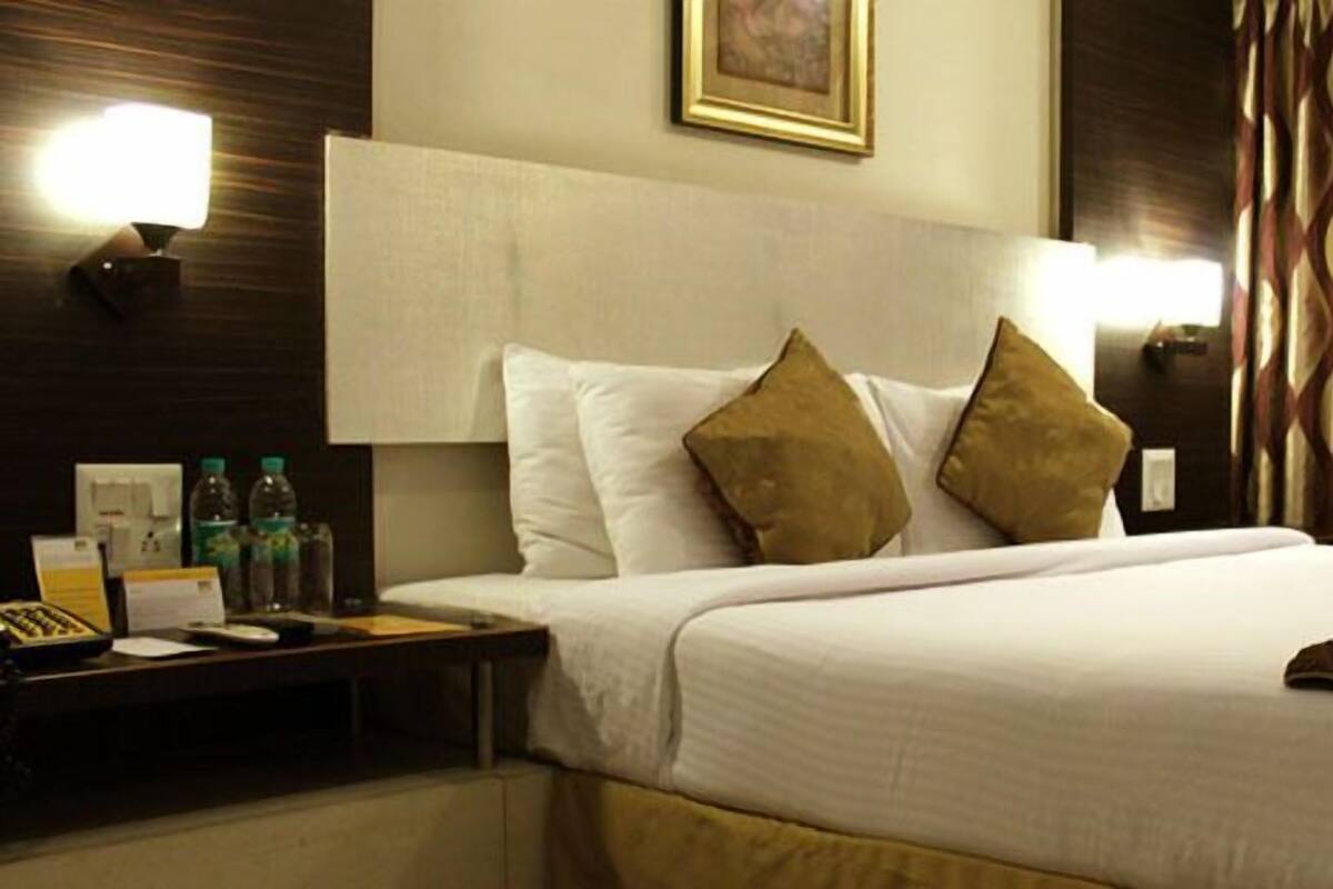 Habitación de hotel con una cama grande, dos almohadas, una mesita de noche con una lámpara, un cuadro en la pared y cortinas estampadas.