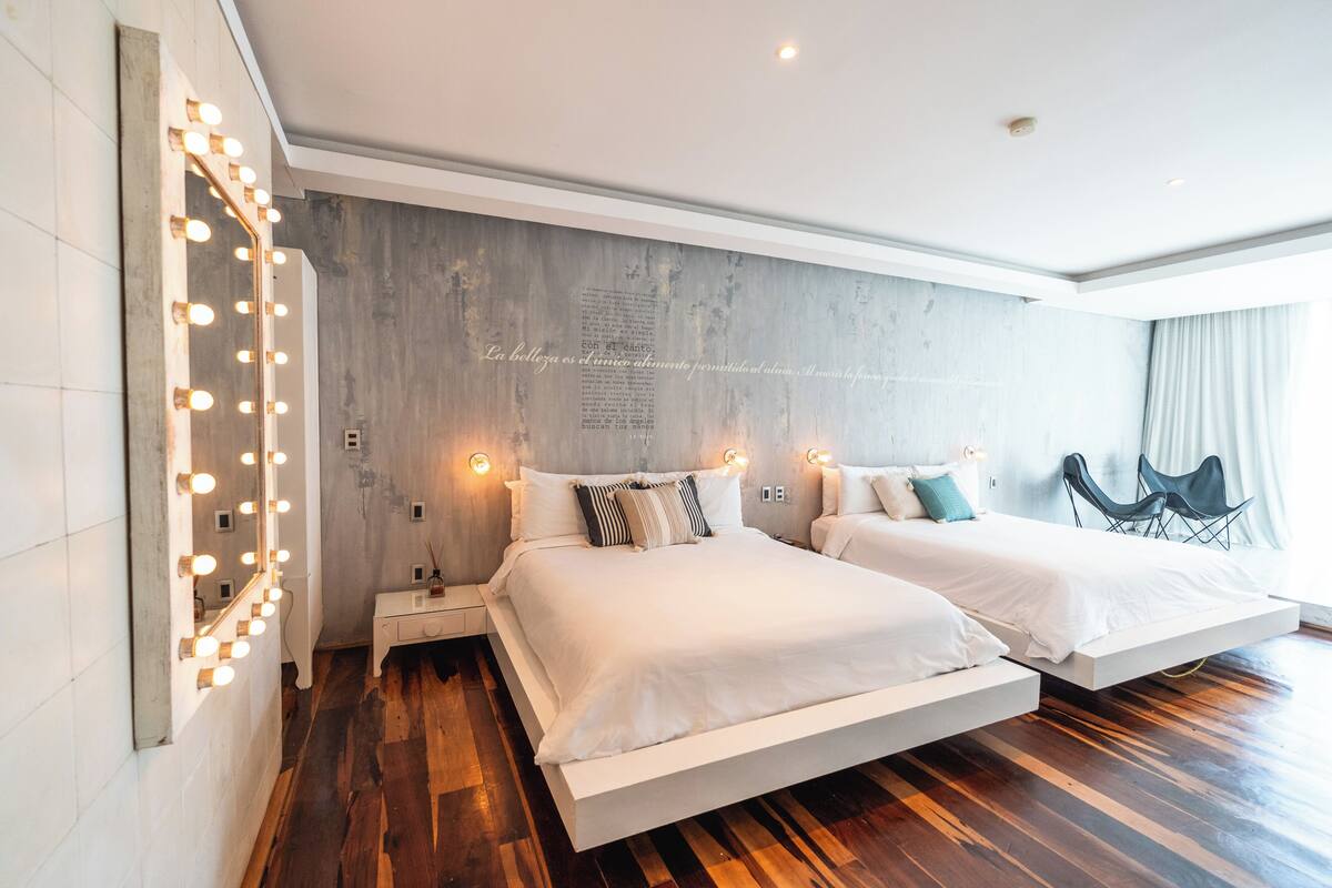 Habitación de hotel moderna con una cama grande, mesitas de noche, tocador con iluminación y vistas al exterior.