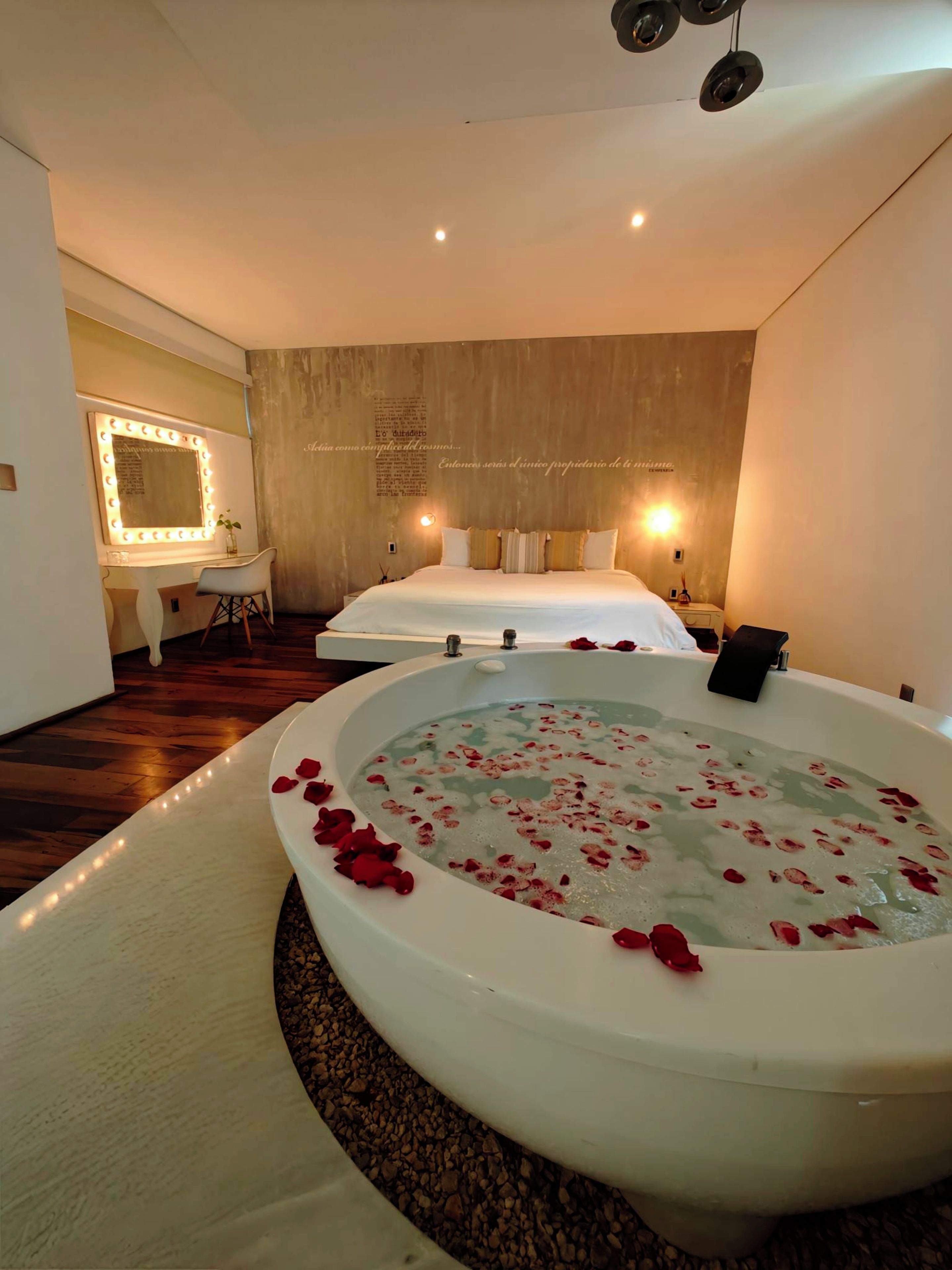 Una habitación con una bañera grande llena de agua y pétalos de rosa, un tocador con espejo e iluminación, y una cama con almohadas.