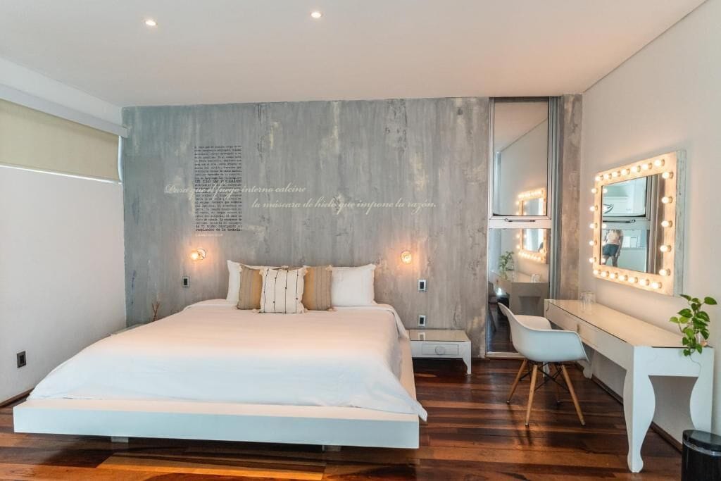 A modern bedroom with a large bed, bedside tables, a vanity with lights, and a mirror.