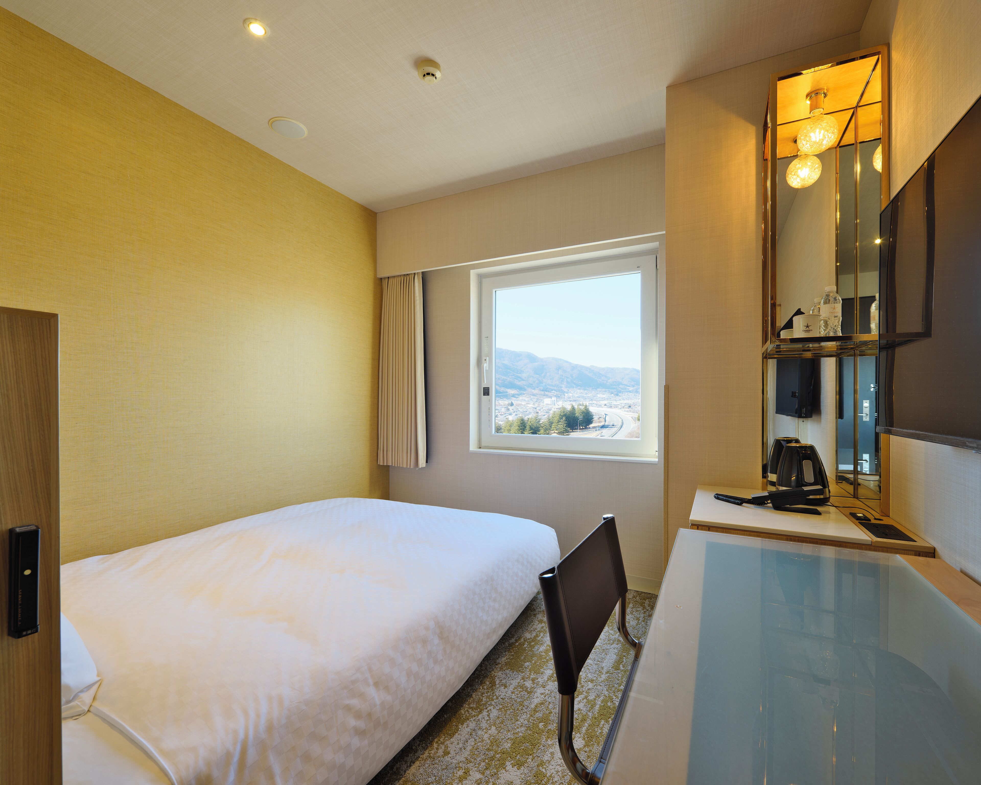 A hotel room with a bed, a desk, a chair, a window with a view, and a mirror.