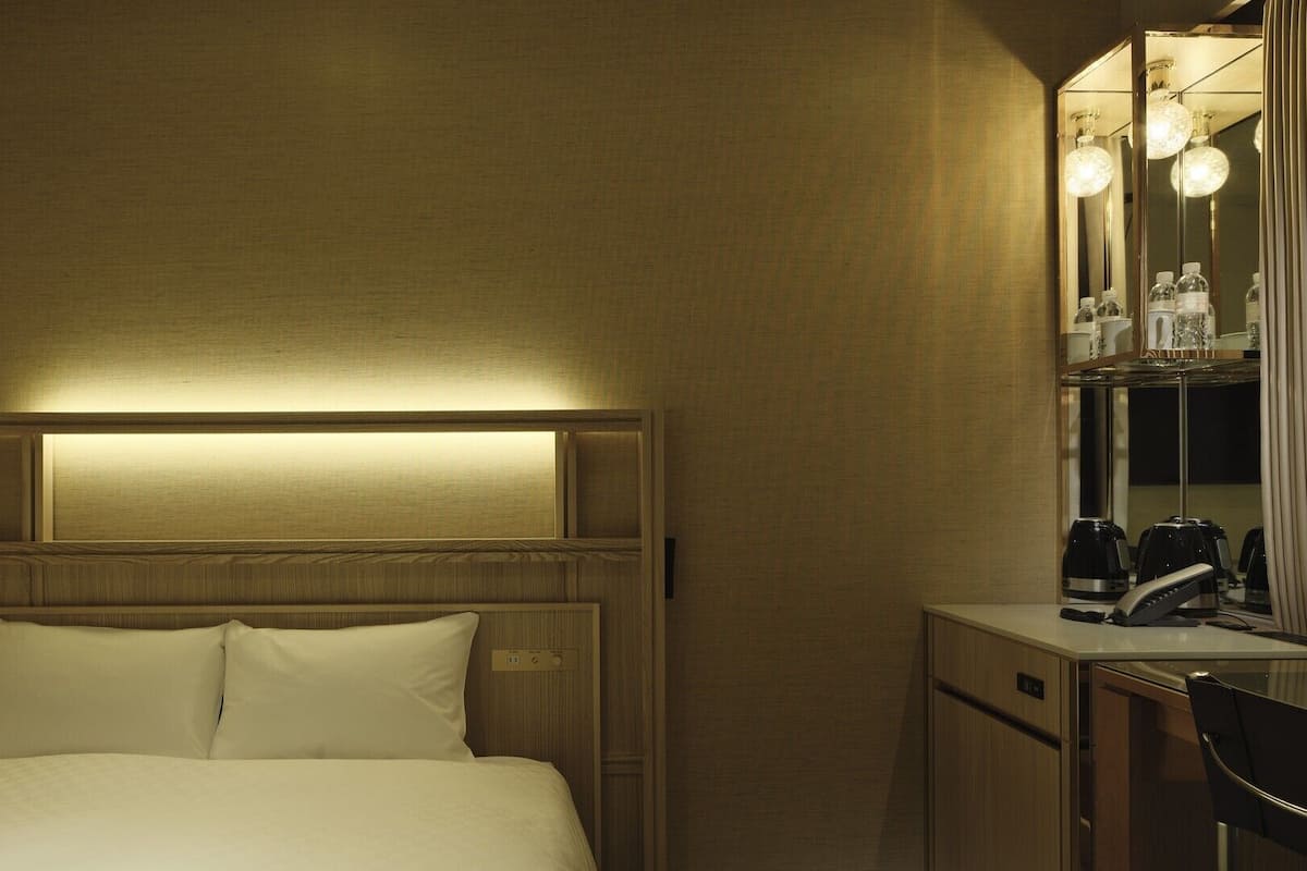 A hotel room with a bed, bedside table, and a mirror with lights.