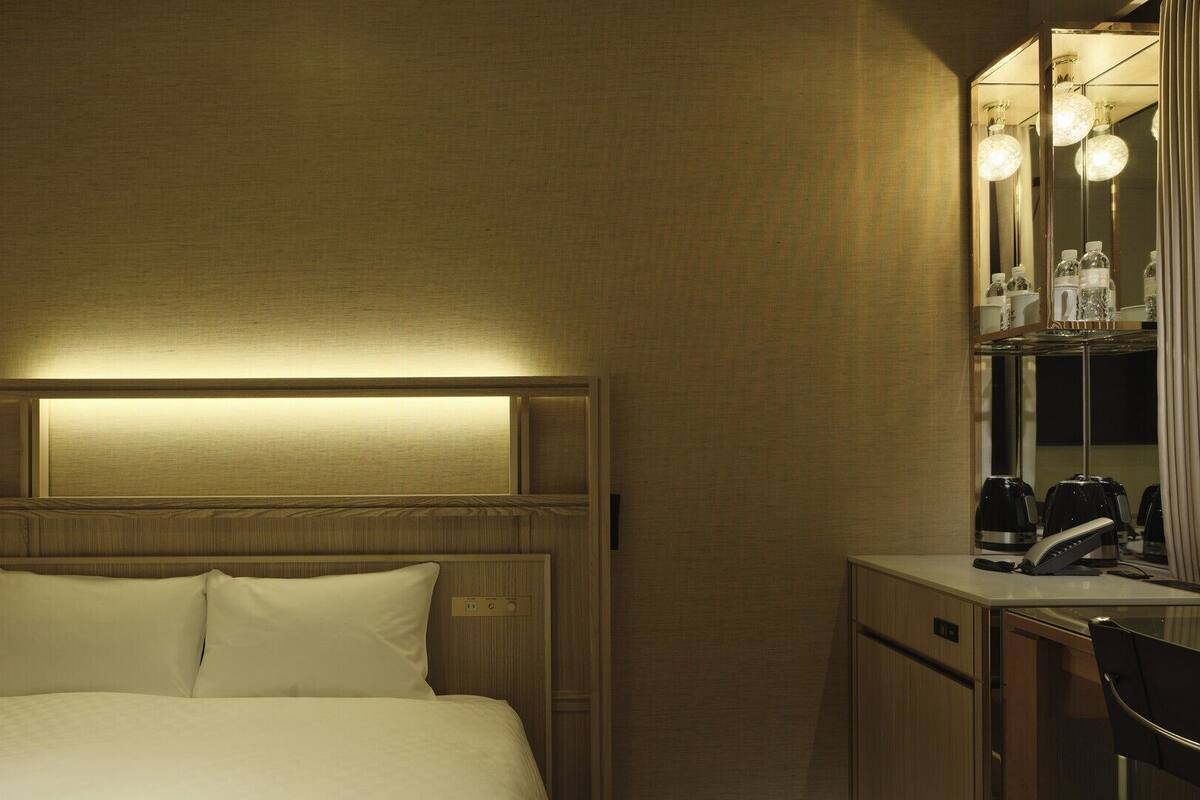 A hotel room with a bed, bedside table, and a mirror with lights.