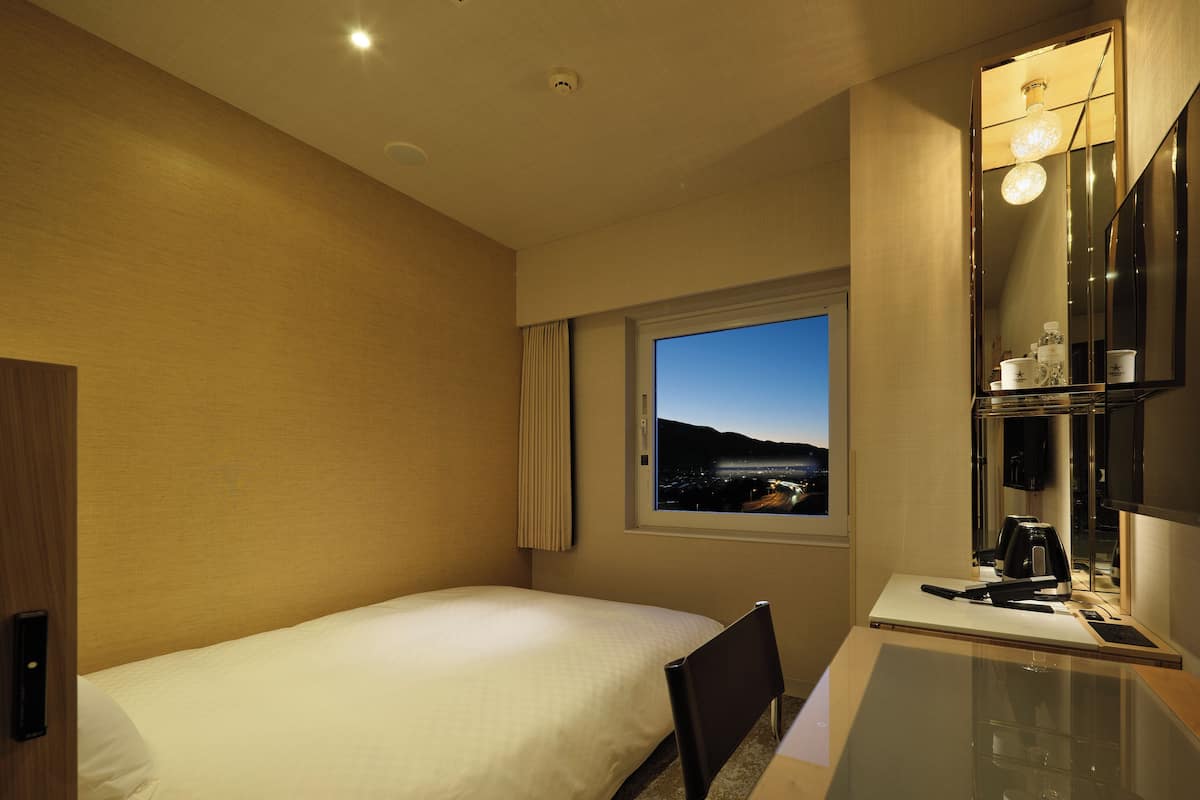 A hotel room with a bed, a desk with a laptop, a chair, a window with a city view, and a mirror.