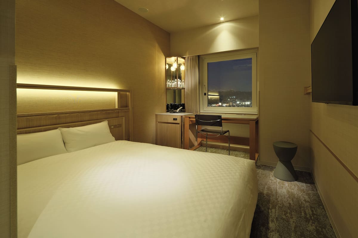 A hotel room with a bed, desk, chair, and a window with a city view at night.