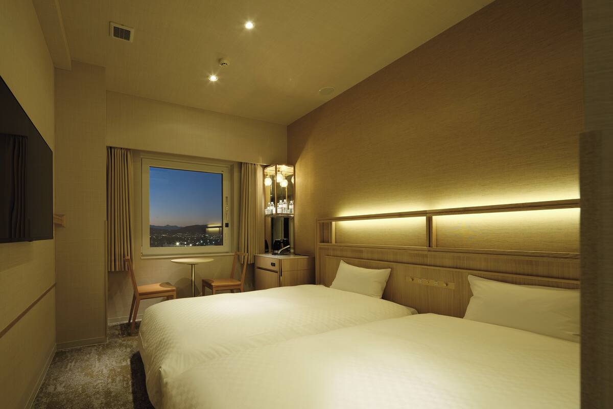 A hotel room with a large bed, a desk, a chair, a mirror, and a window with a city view.