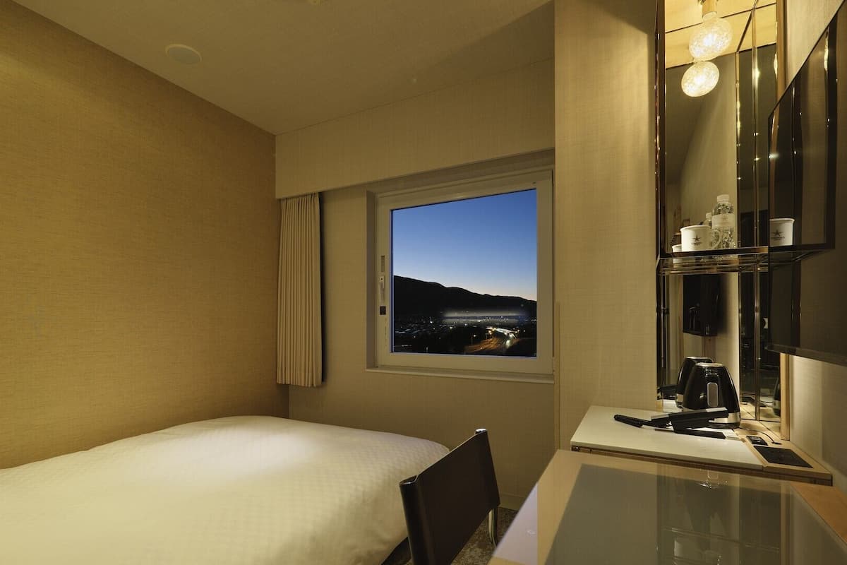 A hotel room with a bed, a desk with a computer, a window with a view of a landscape, and a mirror.