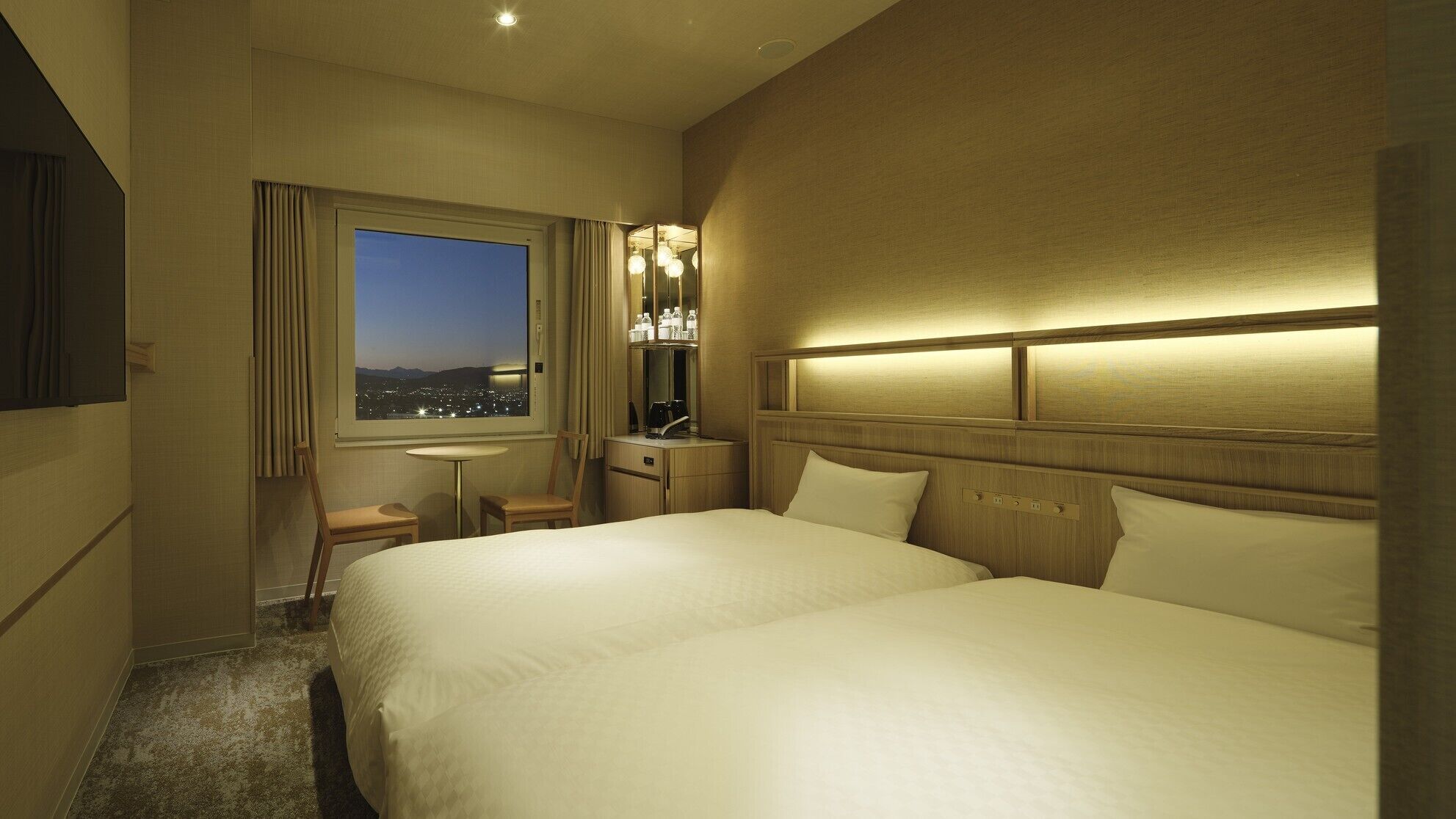 Une chambre d’hôtel avec deux lits, un bureau, une chaise, une fenêtre donnant sur la ville et un mini-bar.