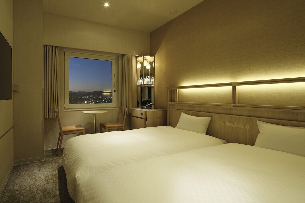 A hotel room with two beds, a desk, a chair, a window with a city view, and a minibar.