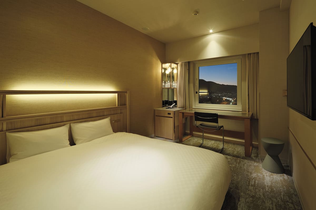 A hotel room with a large bed, a desk with a chair, a window with a city view, and a TV mounted on the wall.