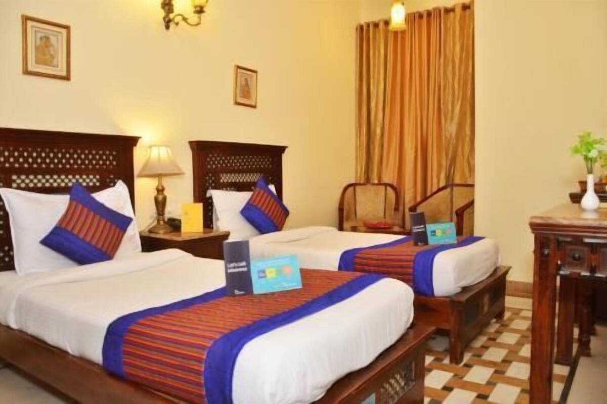 Une chambre d’hôtel avec deux lits, une tête de lit en bois, une table de chevet avec une lampe, une chaise et un bureau agrémenté d’un vase.
