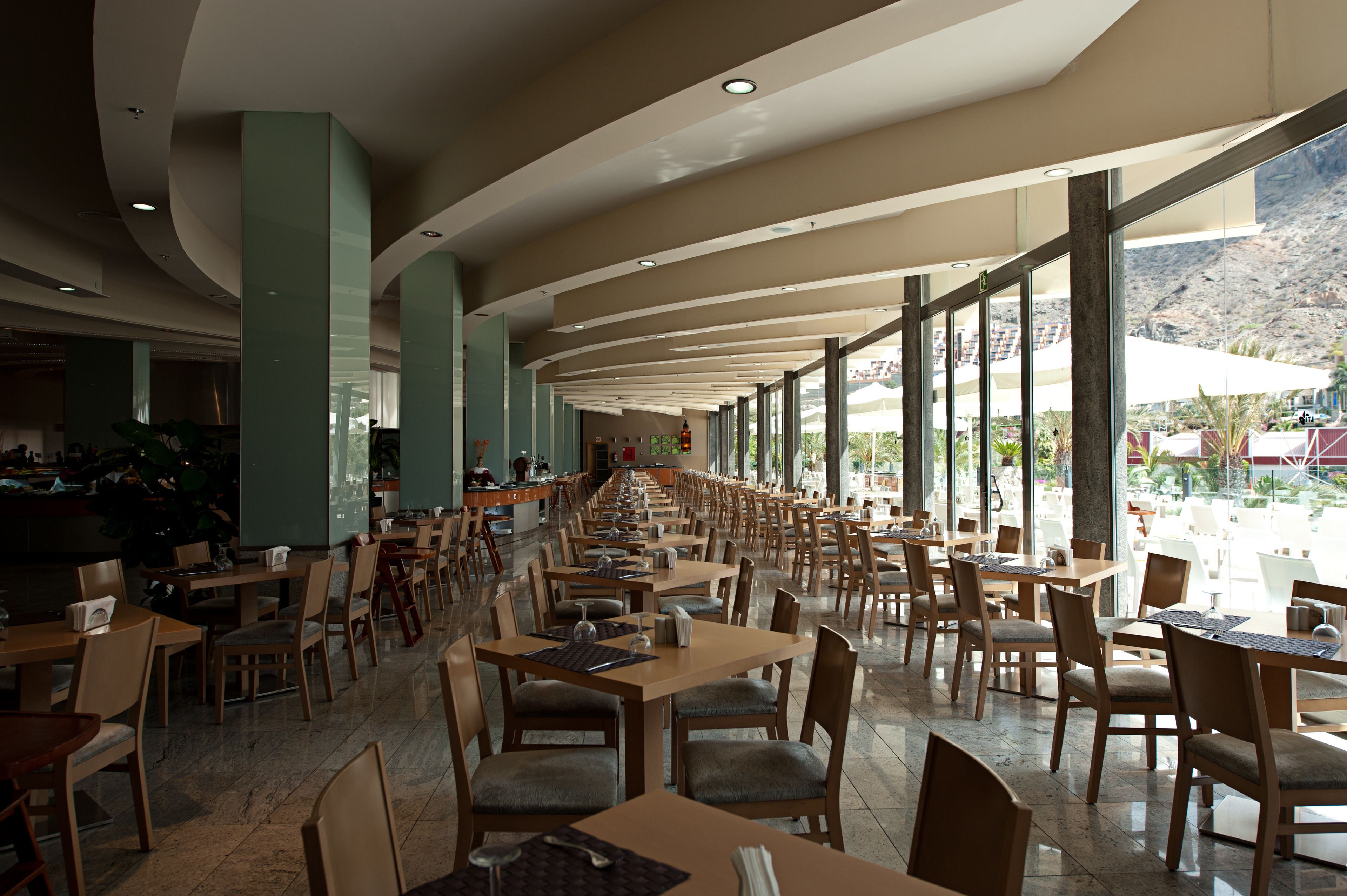 Un'ampia sala da pranzo interna con numerosi tavoli e sedie, ampie finestre che offrono vista sull'esterno e un design del soffitto curvo.