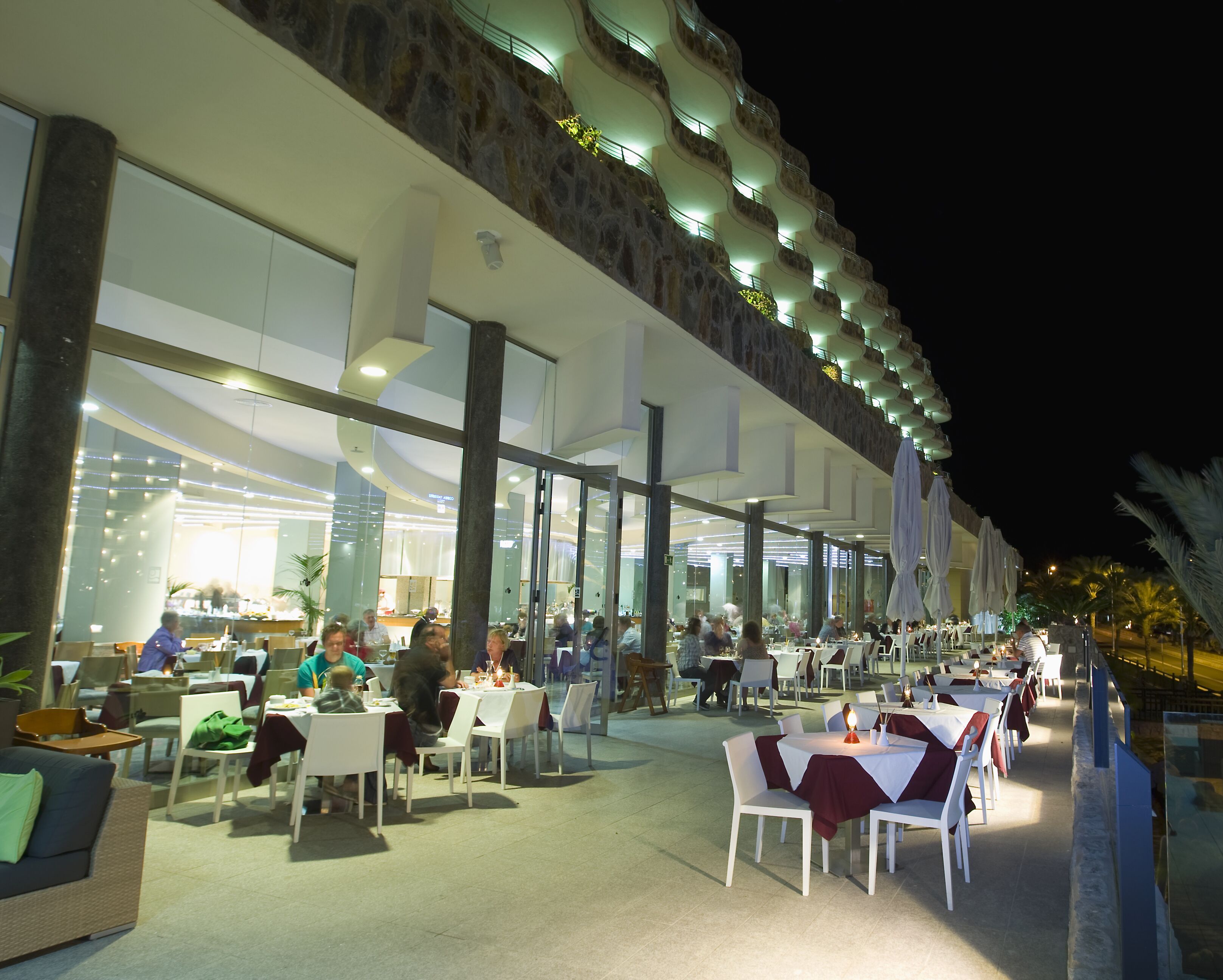 An outdoor dining area with tables and chairs, a stone facade, and a balcony with lights.