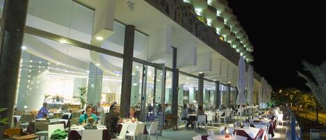 An outdoor dining area with tables and chairs, a stone facade, and a balcony with lights.