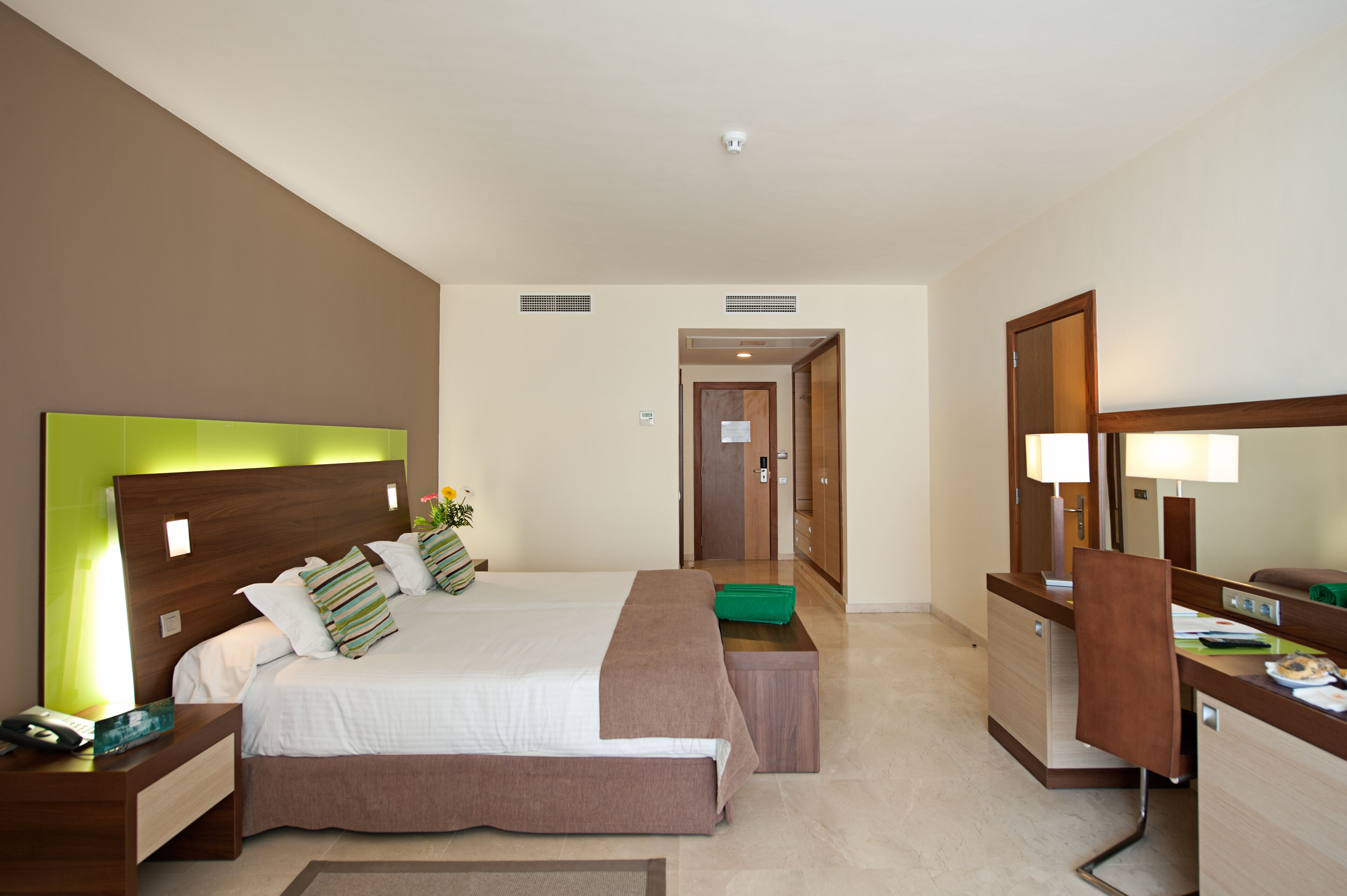A hotel room with a large bed, bedside tables, a desk with a chair, a mirror, and a door leading to another room.