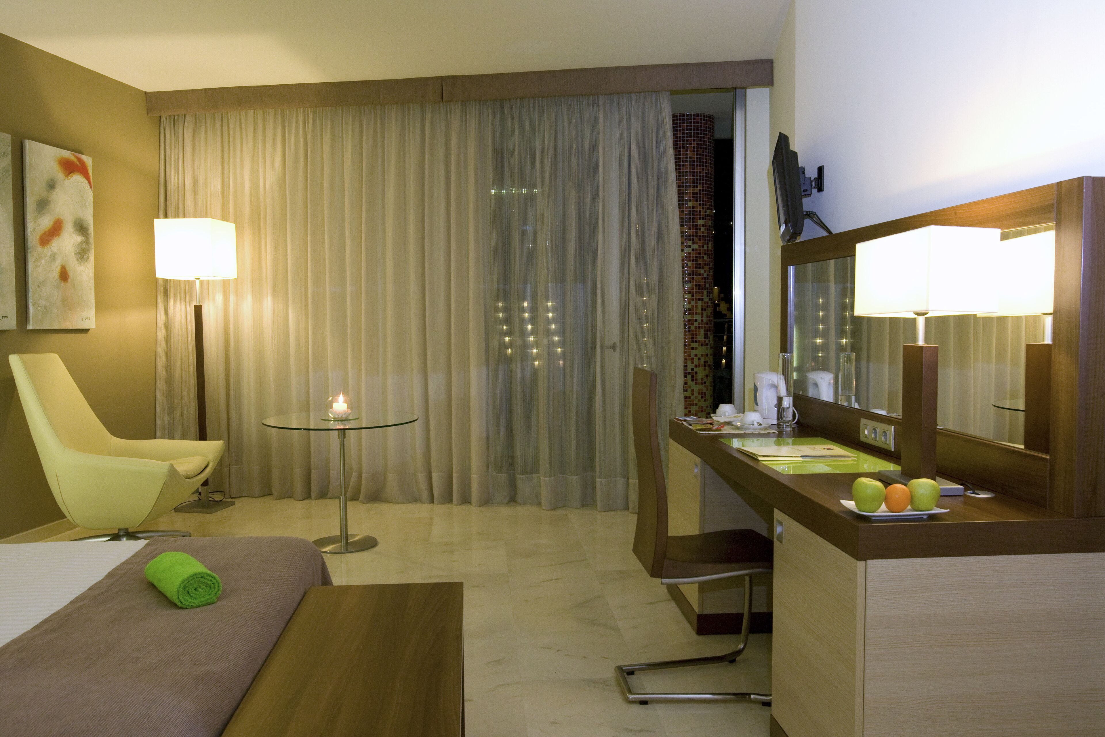 Ein Hotelzimmer mit einem Bett, einem Sessel, einem Schreibtisch mit Spiegel, einem kleinen Tisch mit Lampe und einem Fenster mit Vorhängen.