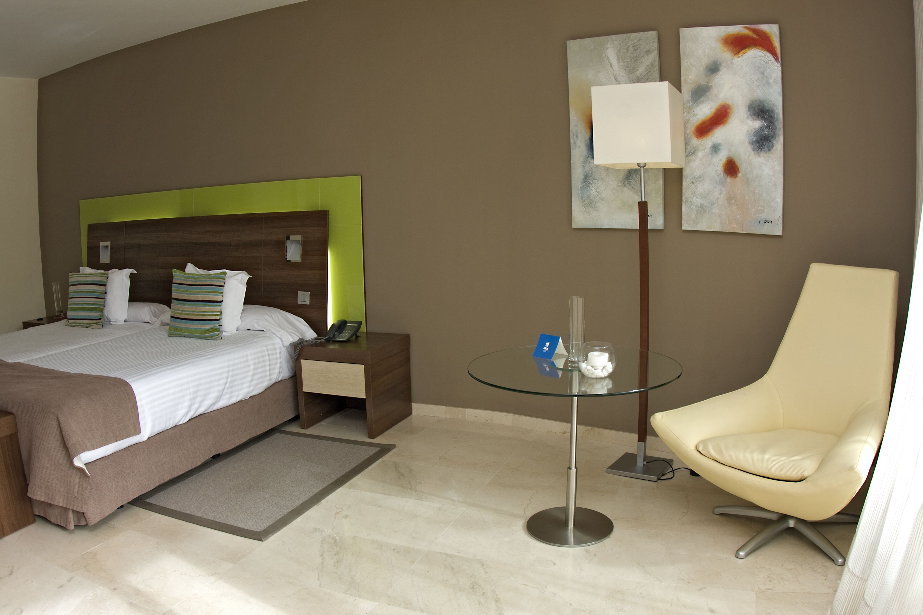 A hotel room with a bed, bedside table, lamp, chair, and artwork on the wall.