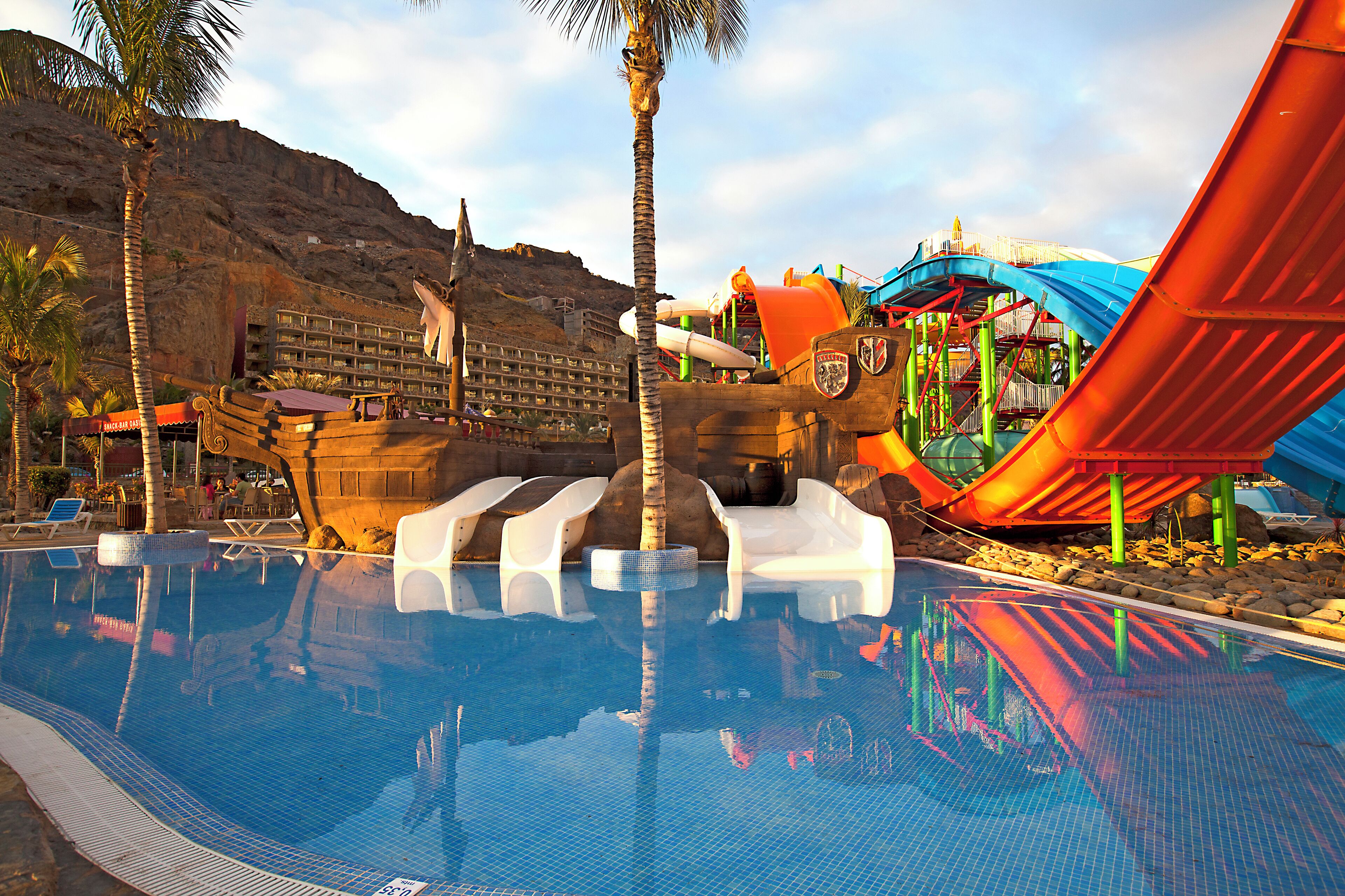 A swimming pool with a water slide and a building with a mountain backdrop.