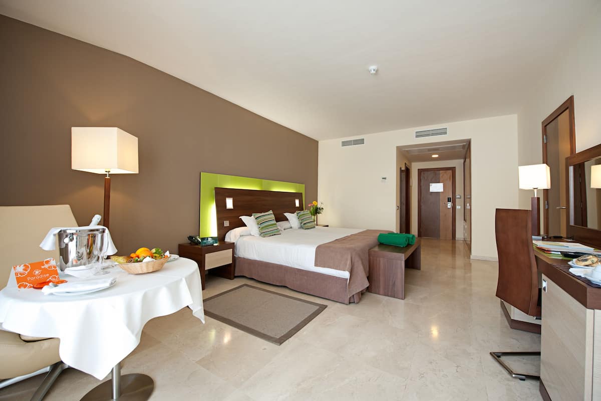 A hotel room with a bed, a desk, a chair, a table with breakfast items, and a bathroom visible in the background.