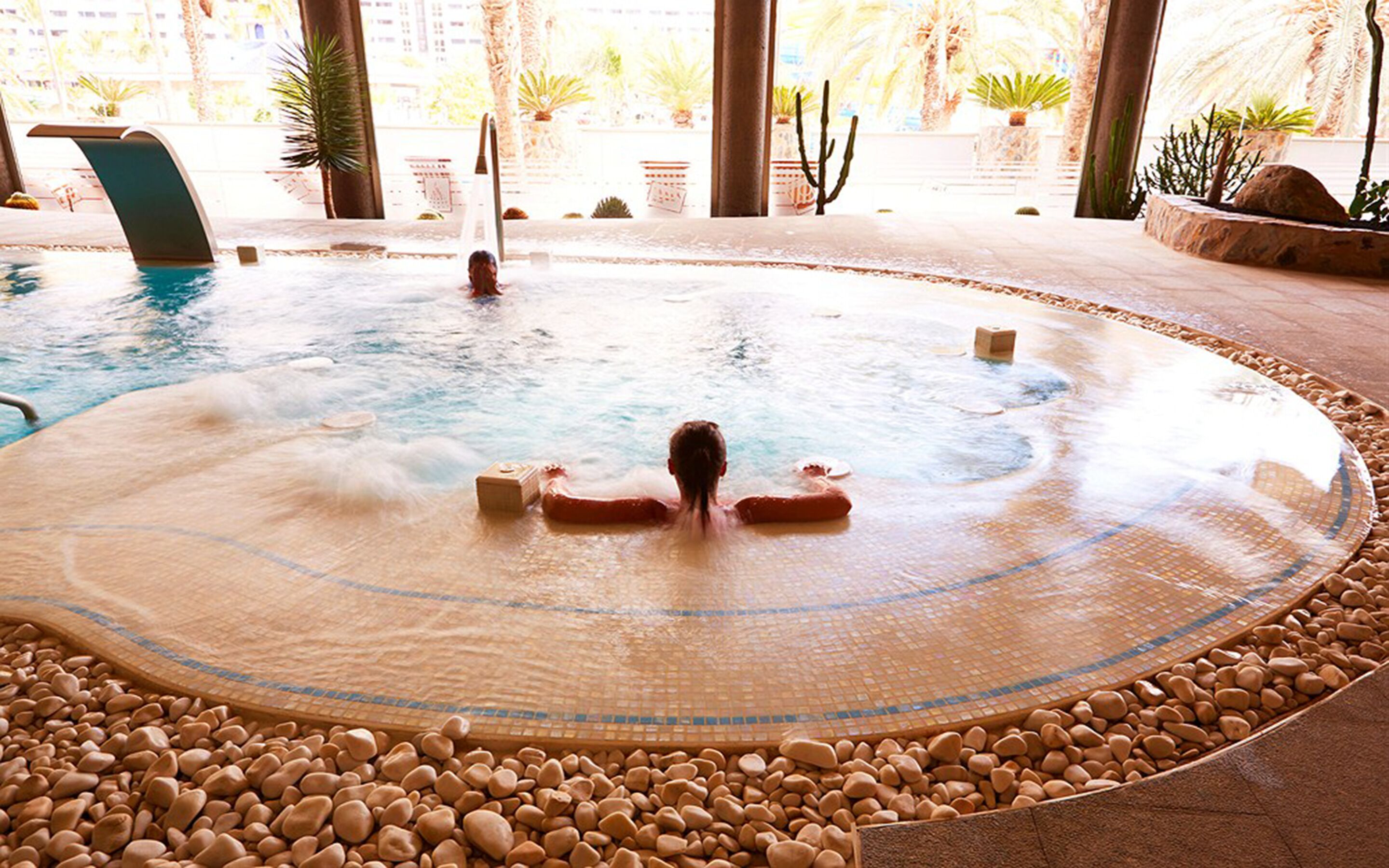 A hot tub with a tiled edge and a pebble border, surrounded by potted plants and palm trees.