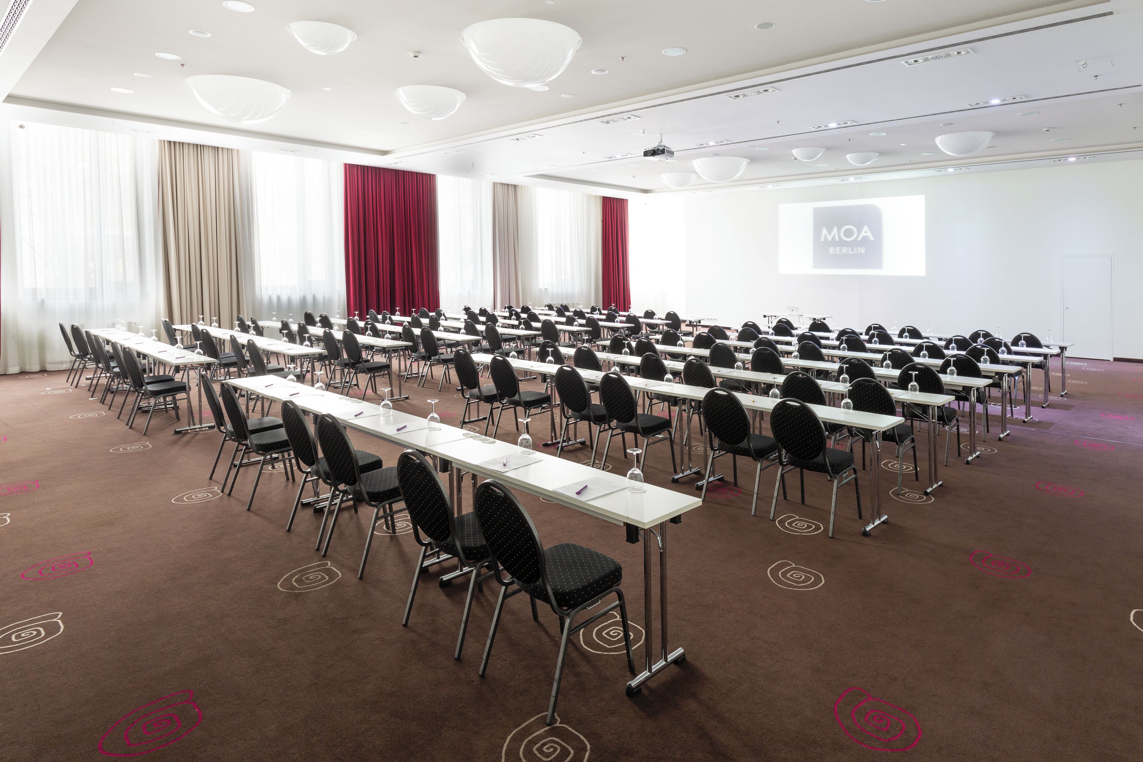 A conference room with rows of tables and chairs, a projector screen displaying 'MOA BERLIN', and a large window with curtains.