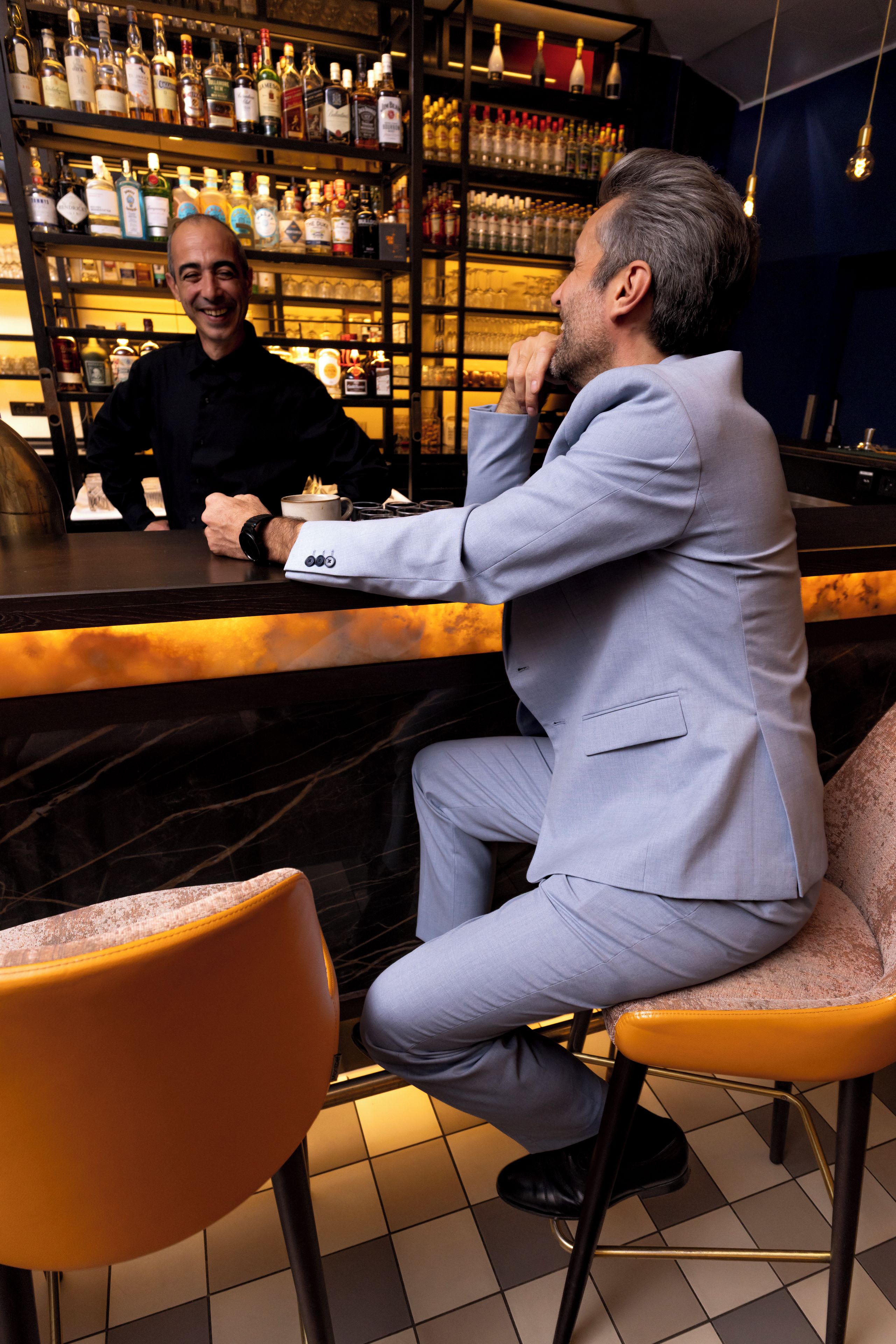 Una persona sentada en un bar, con una estantería repleta de botellas al fondo.
