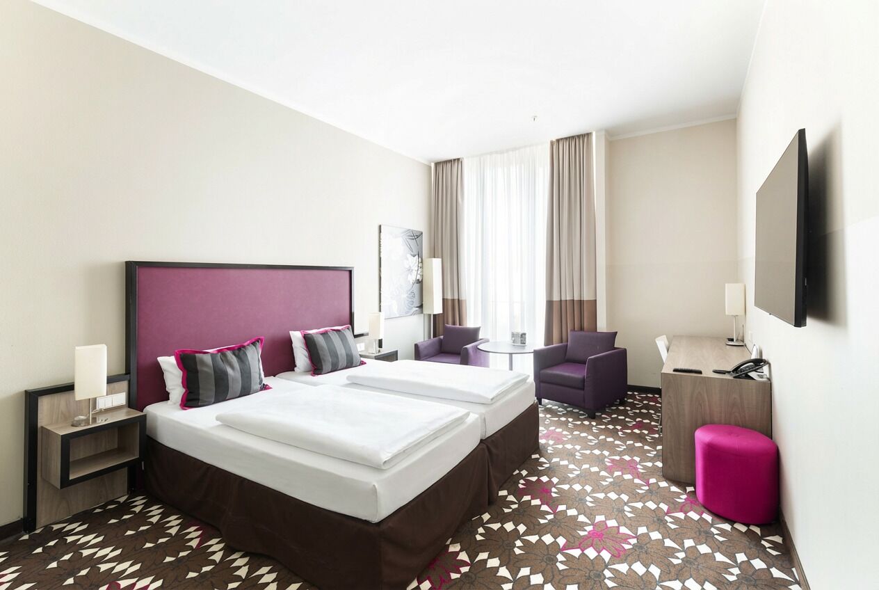 A hotel room with two beds, a desk, a chair, a small table, and a TV.