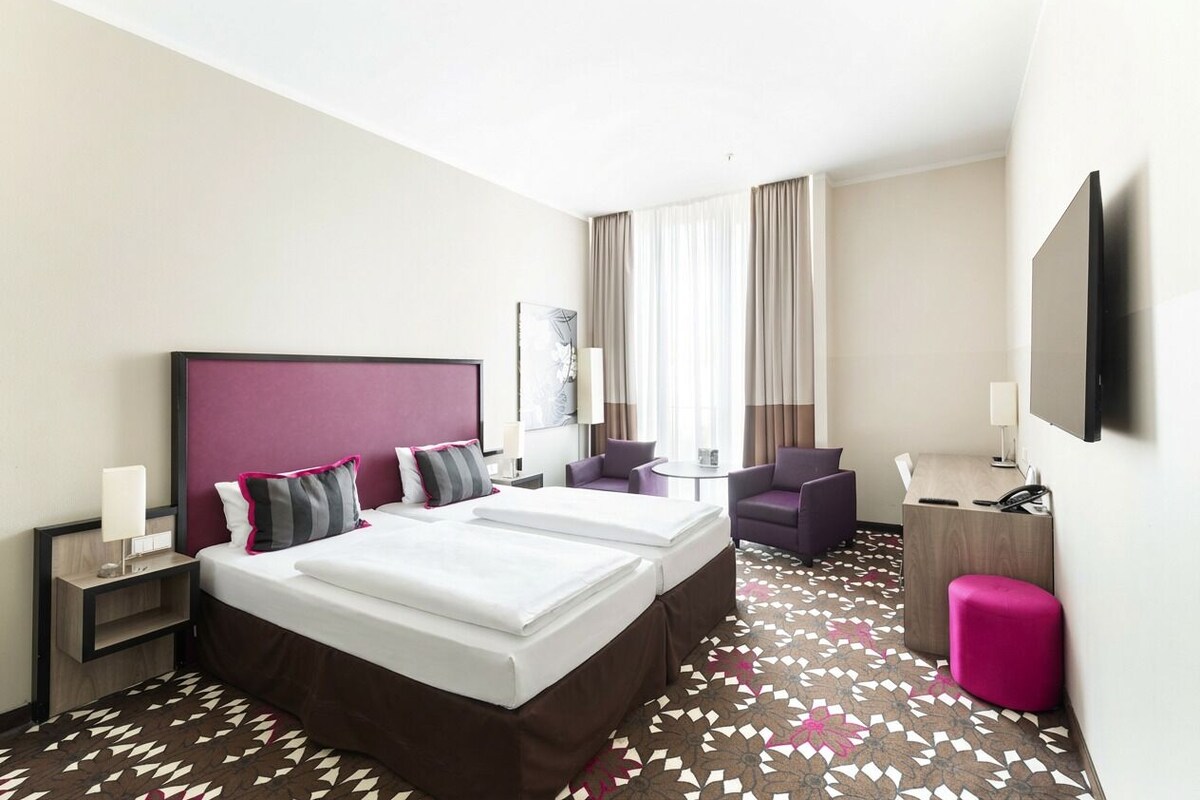 A hotel room with two beds, a desk, a chair, a small table, and a TV.