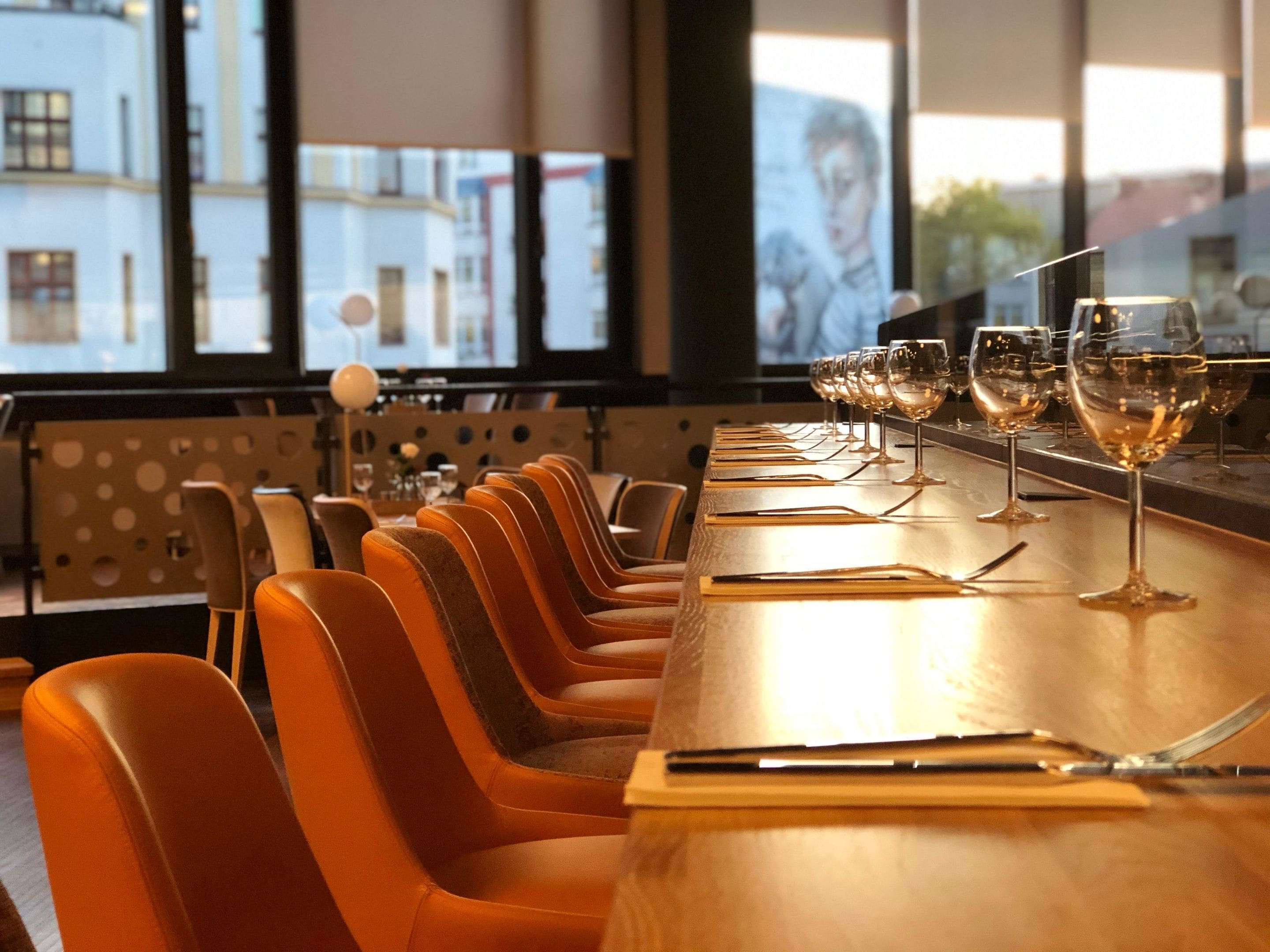 A row of orange chairs and a long table with wine glasses lined up.