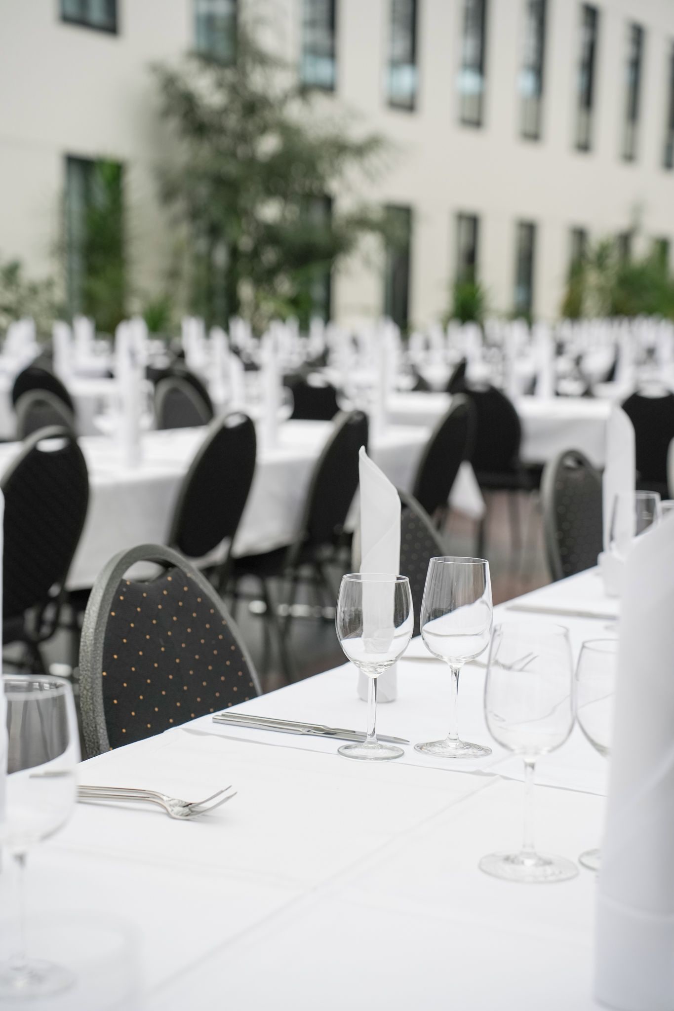 Un'ambientazione da ristorante con tavoli apparecchiati per la cena, caratterizzata da tovaglie bianche, sedie nere e bicchieri trasparenti.
