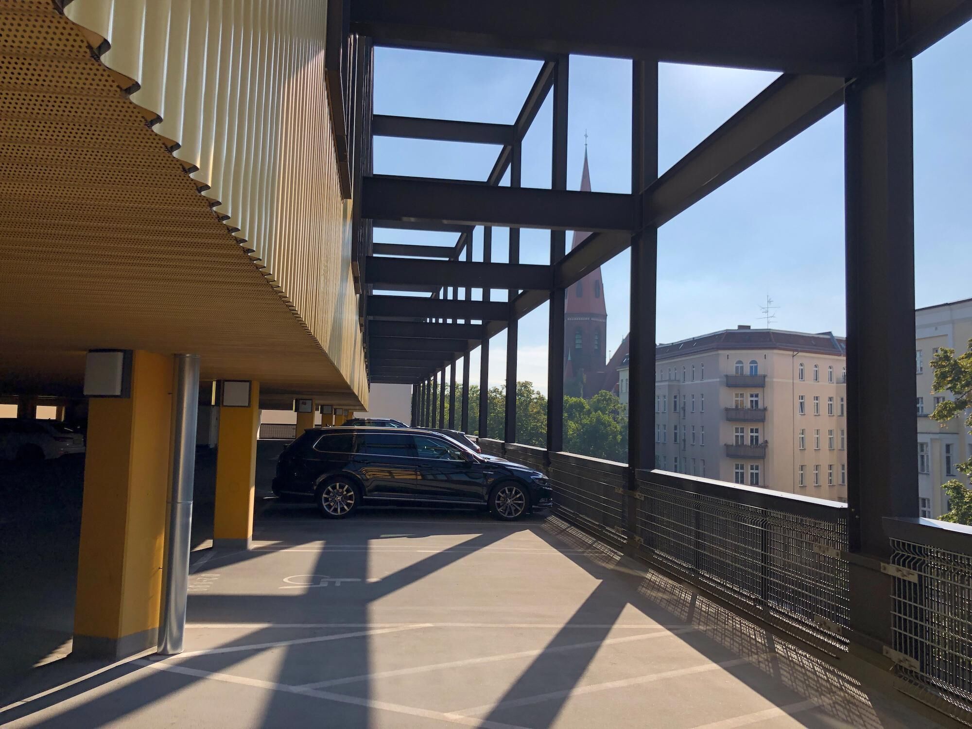Un área de estacionamiento techada con un coche dentro, ventanas amplias y vistas a edificios al fondo.
