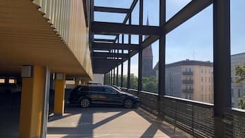Un área de estacionamiento techada con un coche dentro, ventanas amplias y vistas a edificios al fondo.