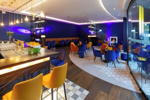 A modern hotel lobby with a bar area, seating with vibrant chairs, and a fireplace.