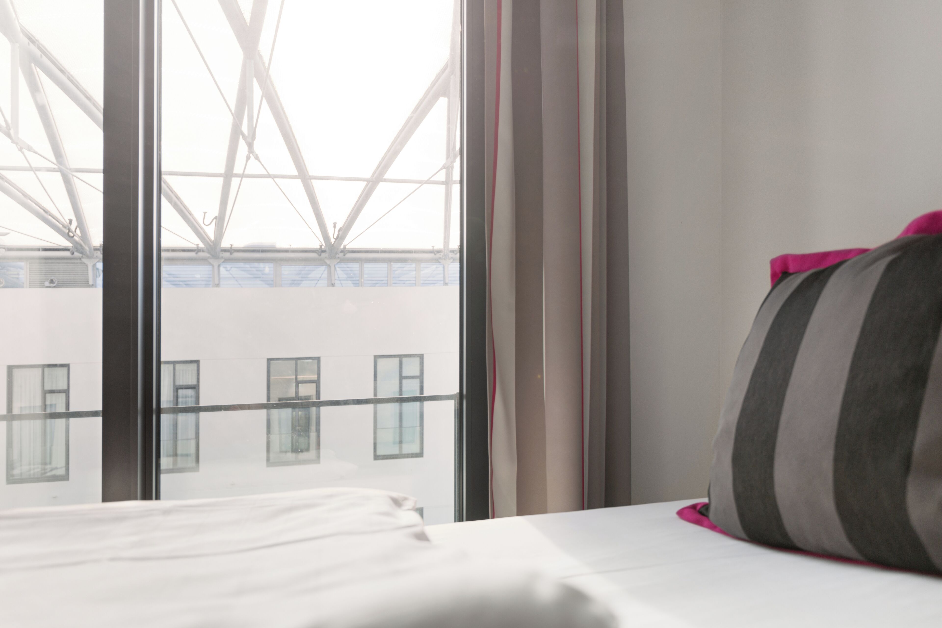 A hotel room with a bed, a window with curtains, and a view of a building with a truss structure.