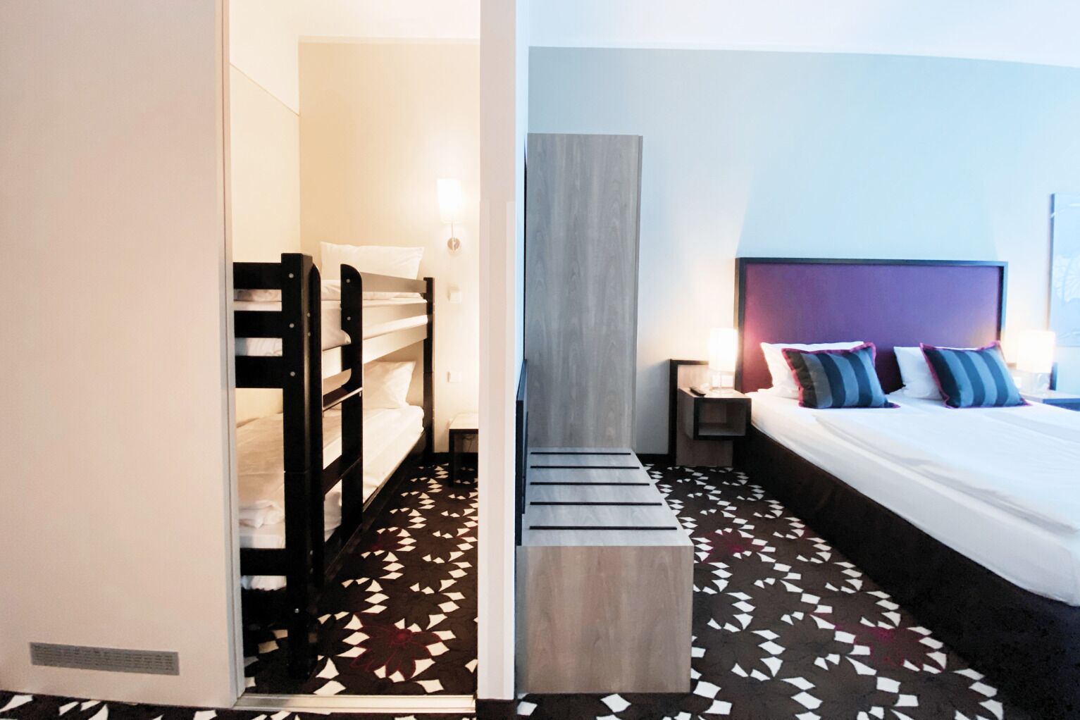 Habitación de hotel con literas, una cama con varias almohadas, una mesita de noche, una lámpara y un suelo con estampado.