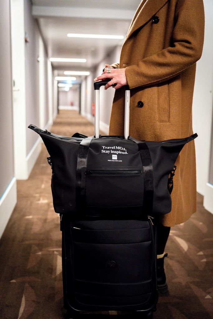 A person in a coat pulling a suitcase with a black bag attached, in a hallway with a carpeted floor and white walls.