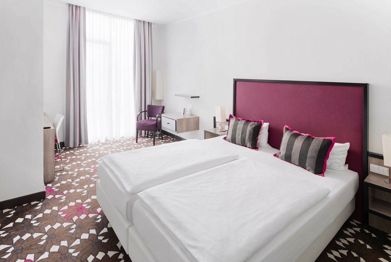 Une chambre d’hôtel avec deux lits, un bureau, une chaise et une grande fenêtre avec des rideaux.