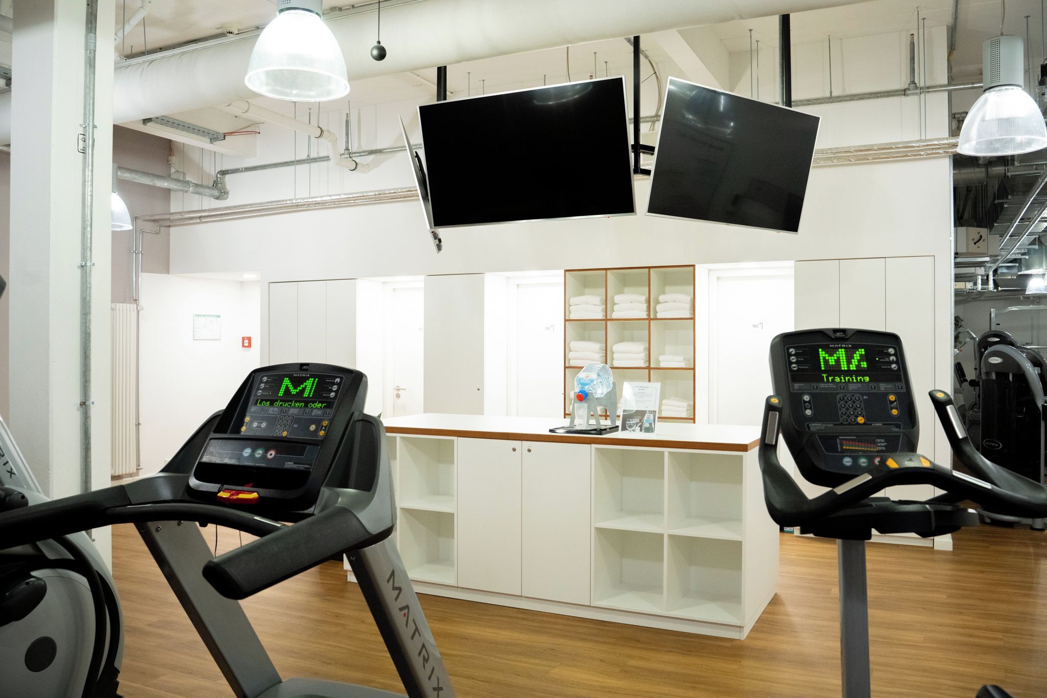 Ein Fitnessraum mit Laufbändern, einem Wasserspender und einem an der Wand befestigten Fernseher.