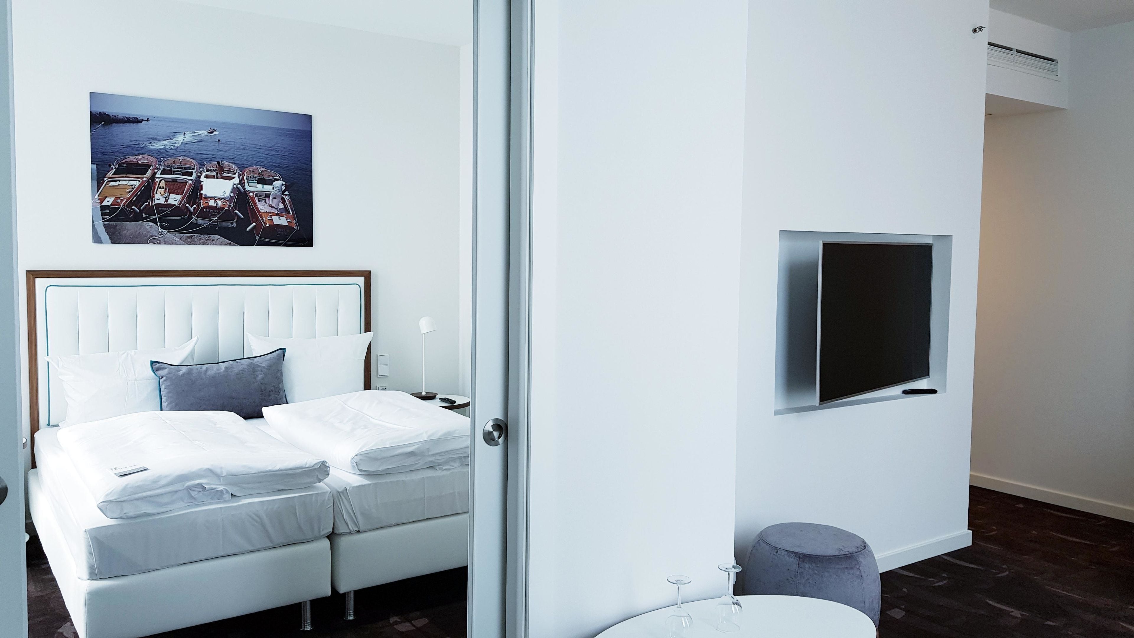 A modern hotel room with a bed, a flat-screen TV mounted on the wall, a small round table with a glass, and a picture of boats on the wall.