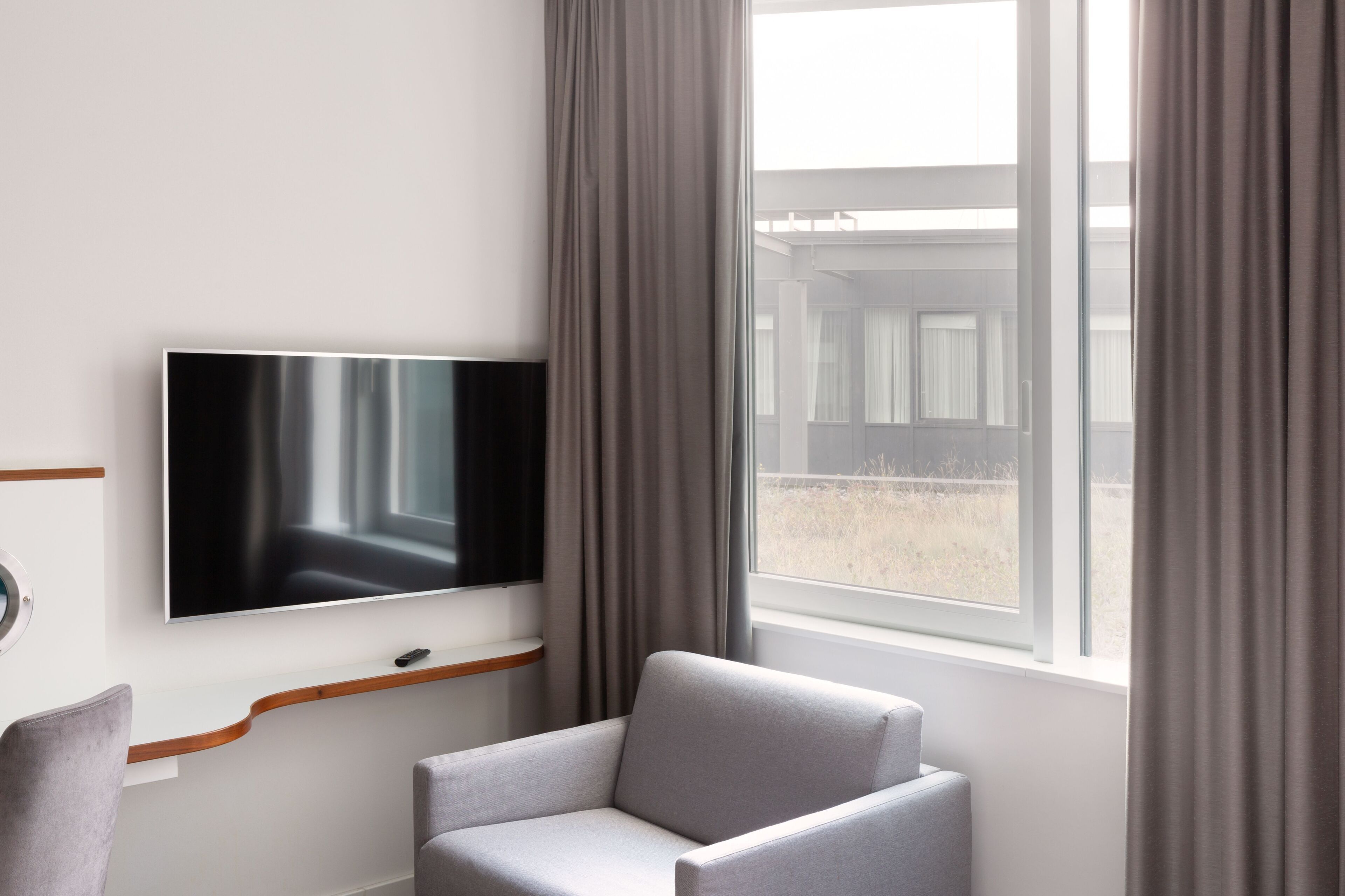A modern room with a flat-screen TV mounted on the wall, a grey armchair, and a window with curtains.