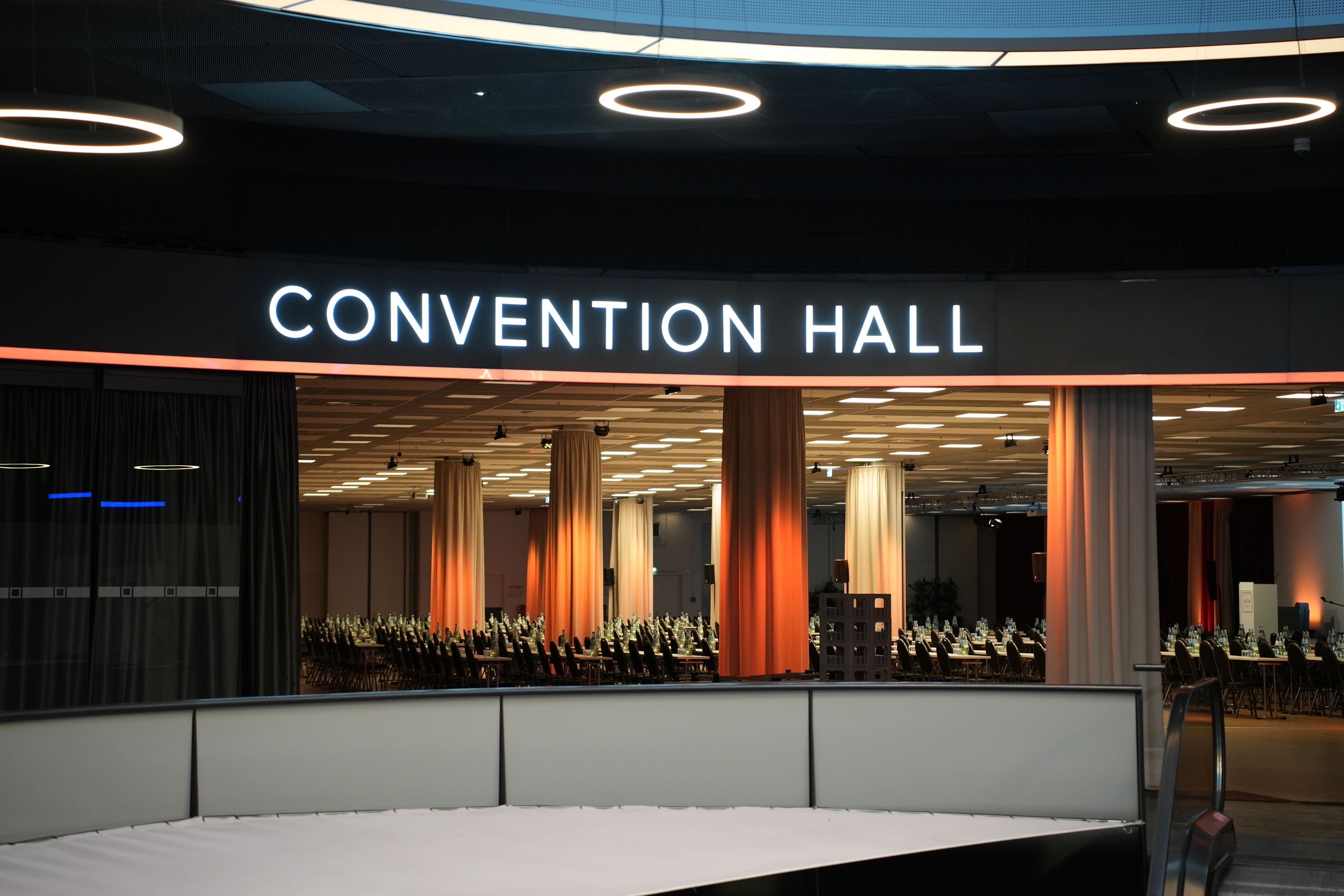 Una sala congressi dal design moderno, con un grande cartello illuminato, soffitti alti e un ampio interno con file di tavoli e sedie.