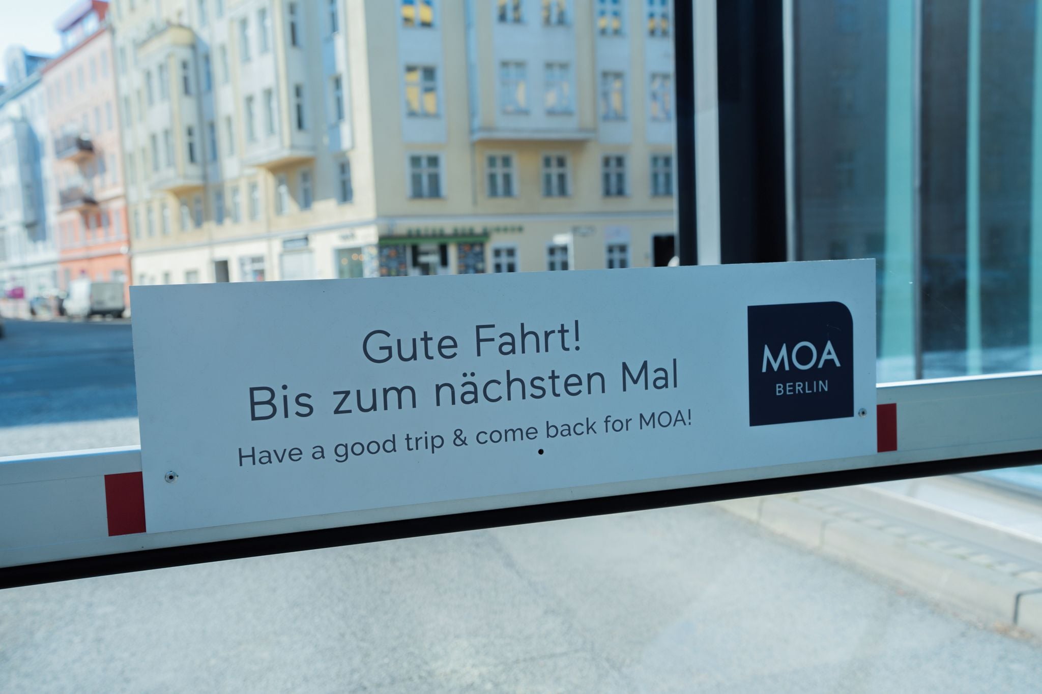 Un cartel con un mensaje en alemán, 'Gute Fahrt! Bis zum nächsten Mal Have a good trip & come back for MOA!' y el logo 'MOA BERLIN' en un edificio.