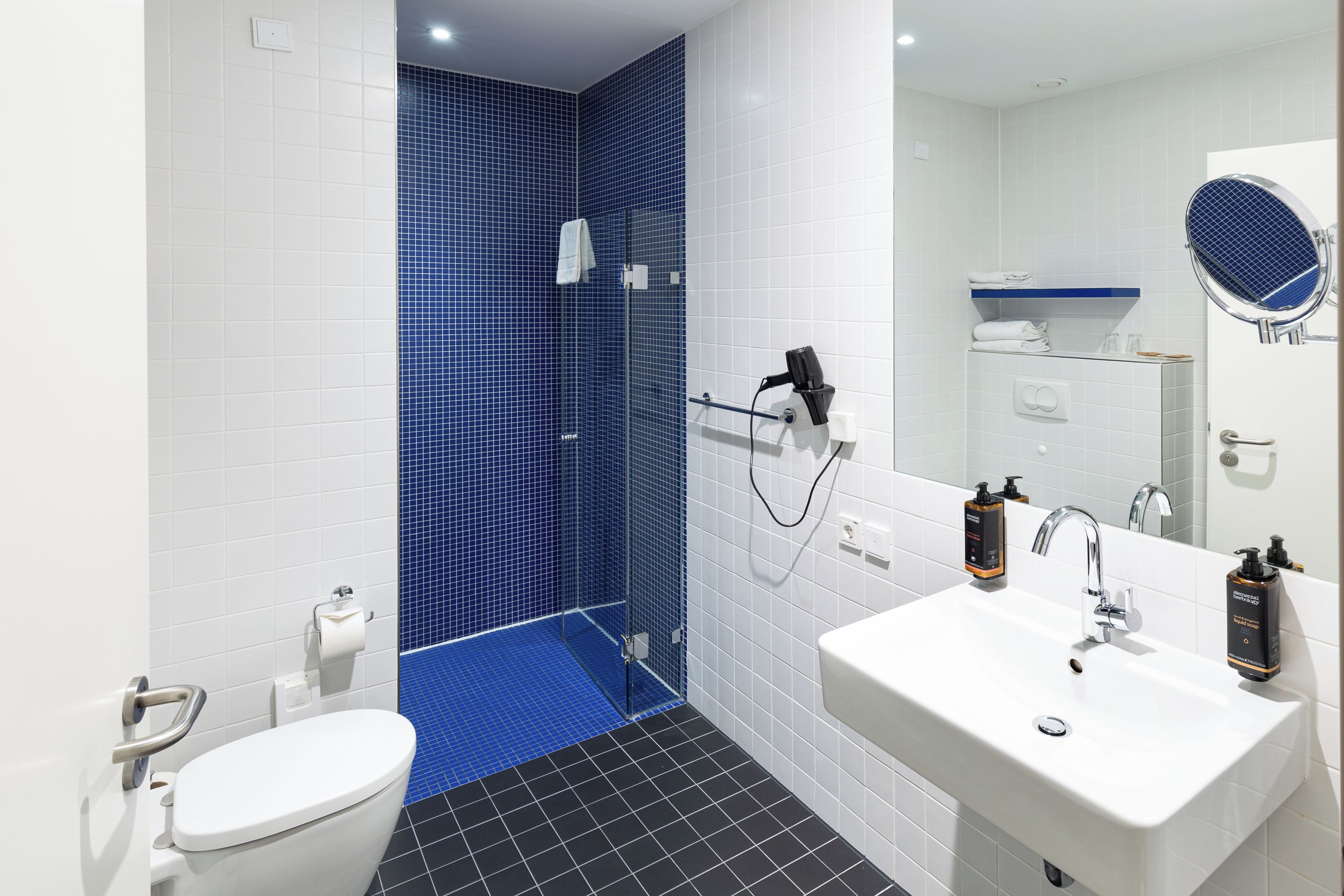 Ein modernes Badezimmer mit Toilette in Weiß, einem Waschbecken mit Spiegel und einer Duschkabine mit blauen Fliesen.