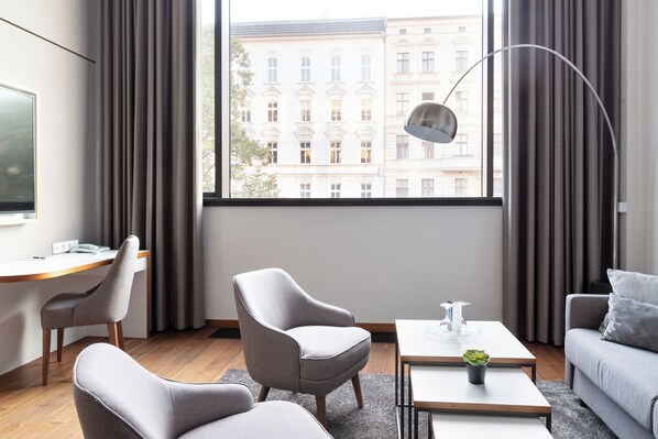 Una habitación de hotel moderna con un escritorio de madera, dos sillones, un sofá, una mesa de centro con una planta y un ventanal con cortinas.