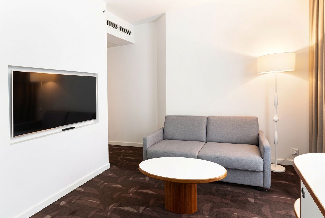 A modern hotel room with a flat-screen TV mounted on the wall, a grey sofa, a round wooden coffee table, and a floor lamp.