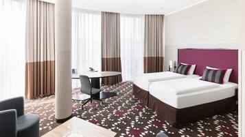 Habitación de hotel con cama, dos mesitas de noche, un escritorio, una silla y un ventanal con cortinas.