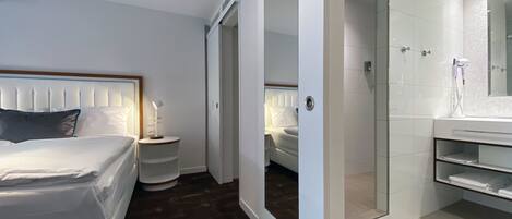 A modern hotel room with a neatly made bed, a bedside table with a lamp, a walk-in closet, and a bathroom with a shower and a large mirror.