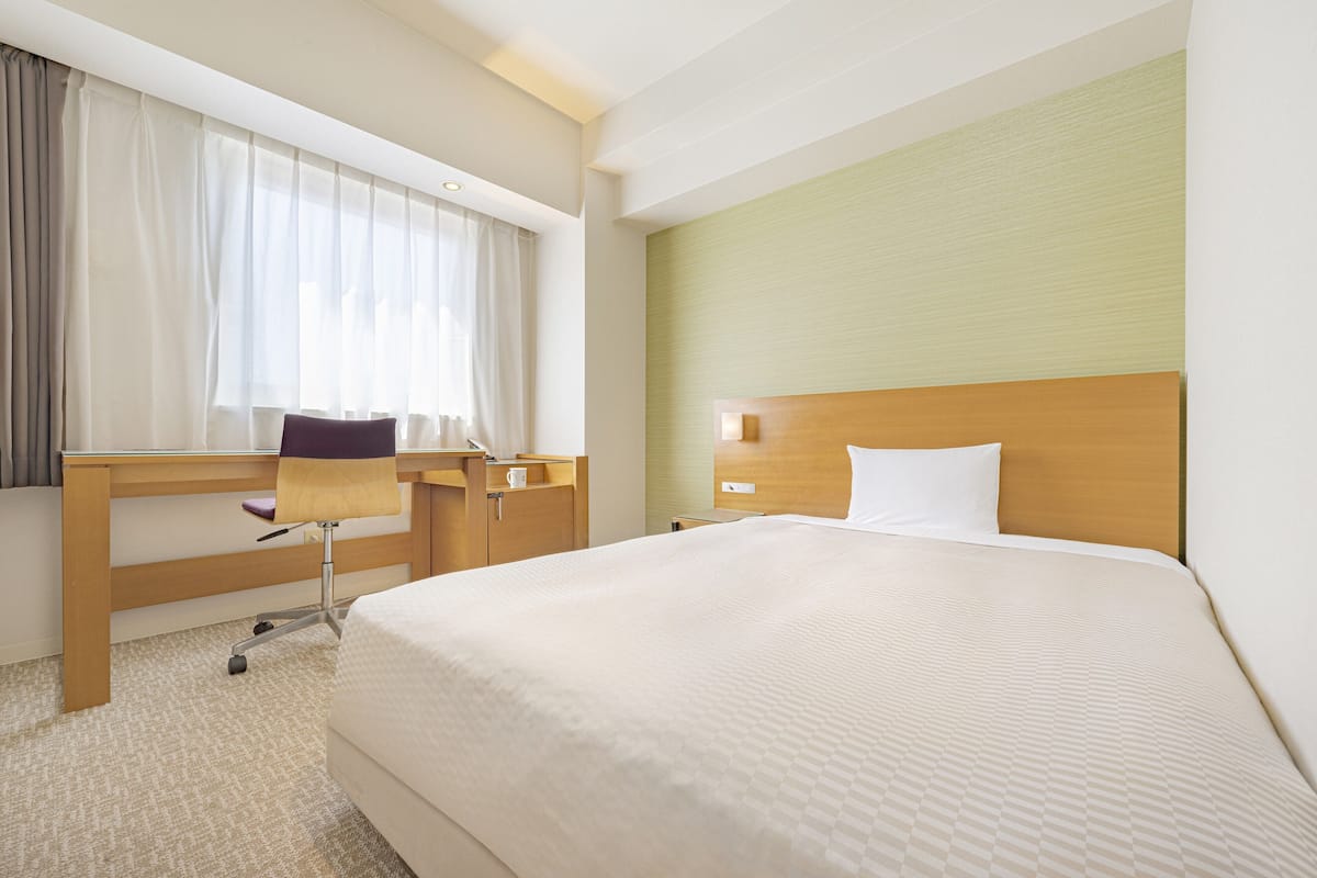 Een hotelkamer met een groot bed, een bureau met stoel, een raam met gordijnen en een hoofdeinde.