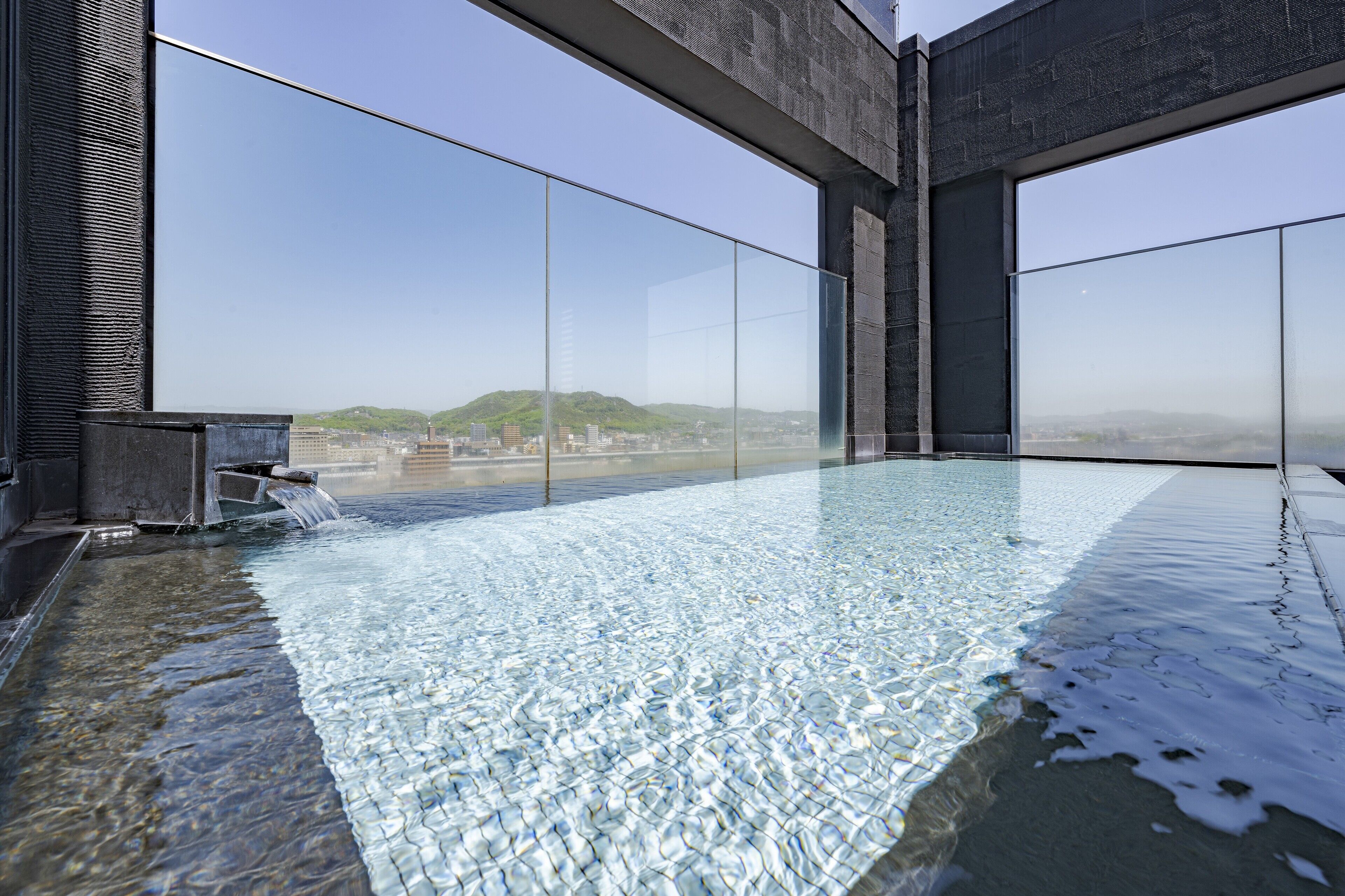 An indoor infinity pool with clear water, surrounded by large windows offering a view of distant hills and buildings.